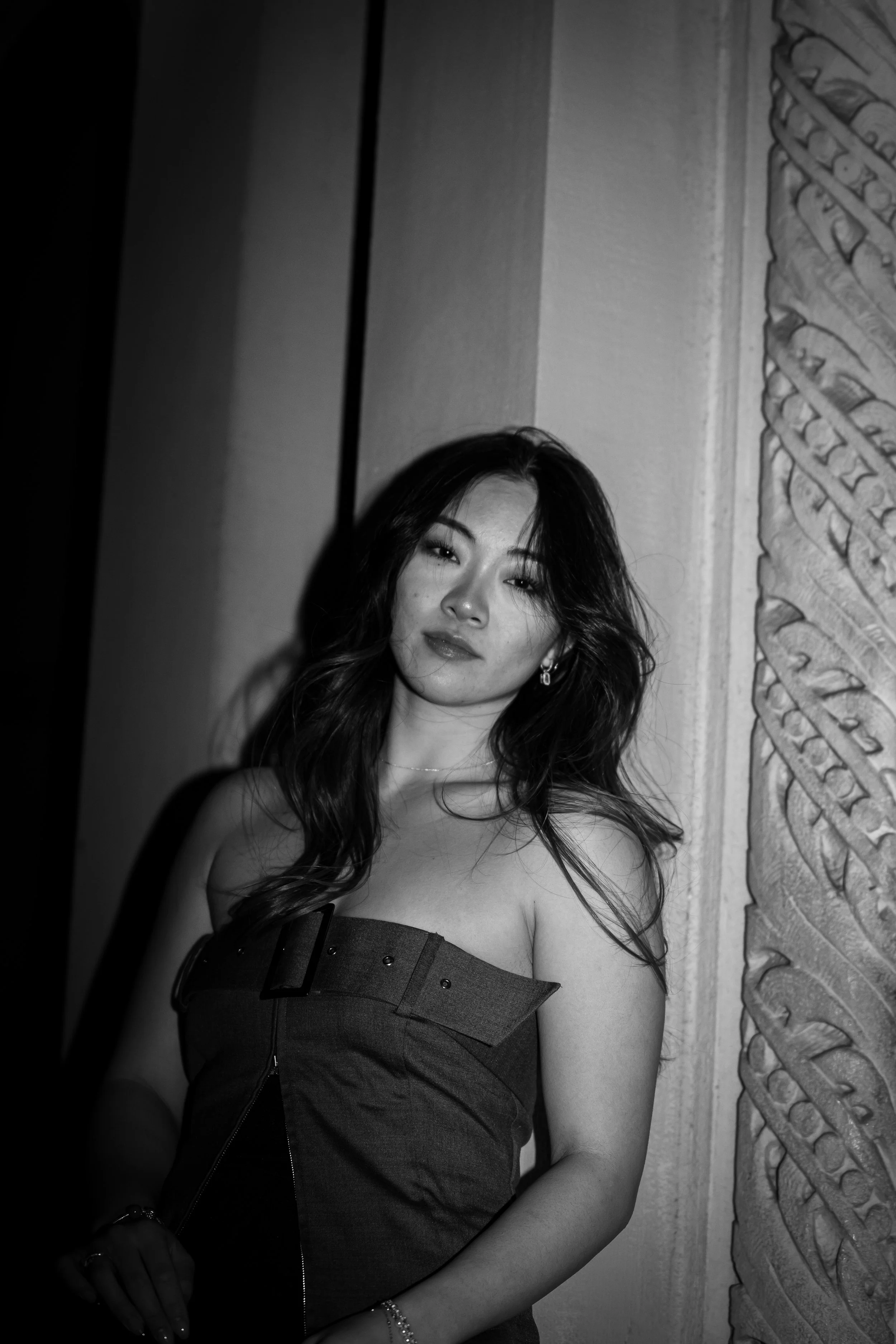 Black and white photo of a young woman with long, wavy dark hair, wearing a strapless dress, standing next to a decorative wall, looking at the camera.