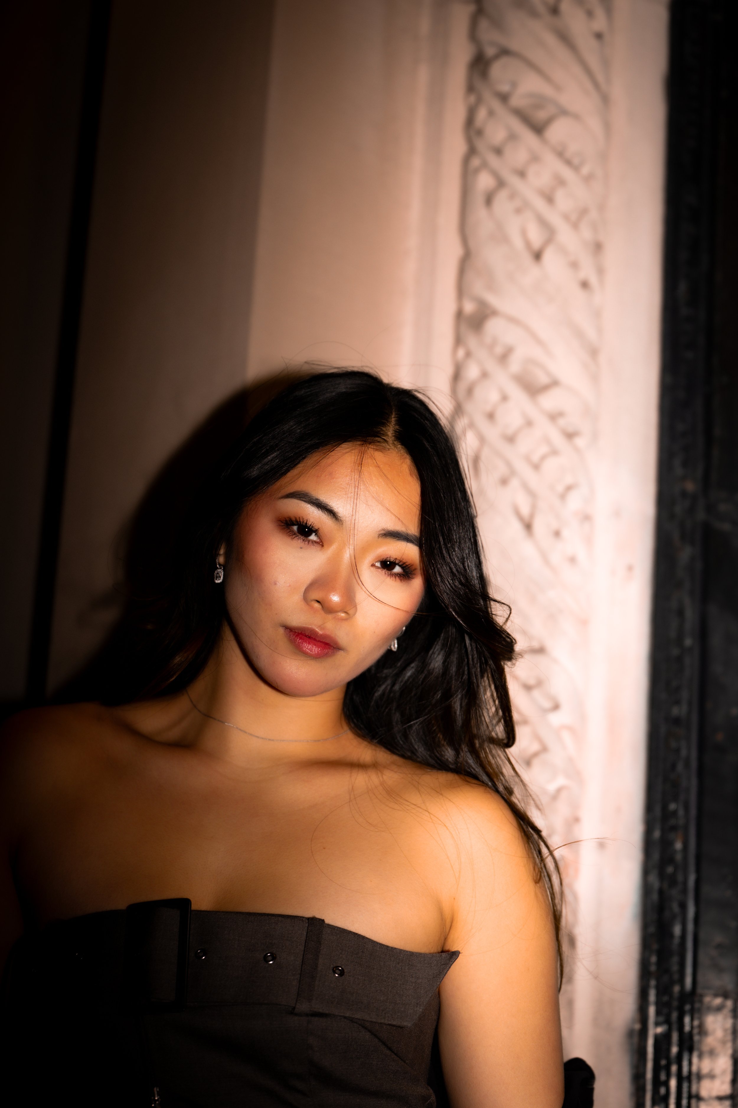 Young woman with long black hair, wearing earrings, necklace and strapless black top, standing against a stone wall with ornate carvings, illuminated softly.