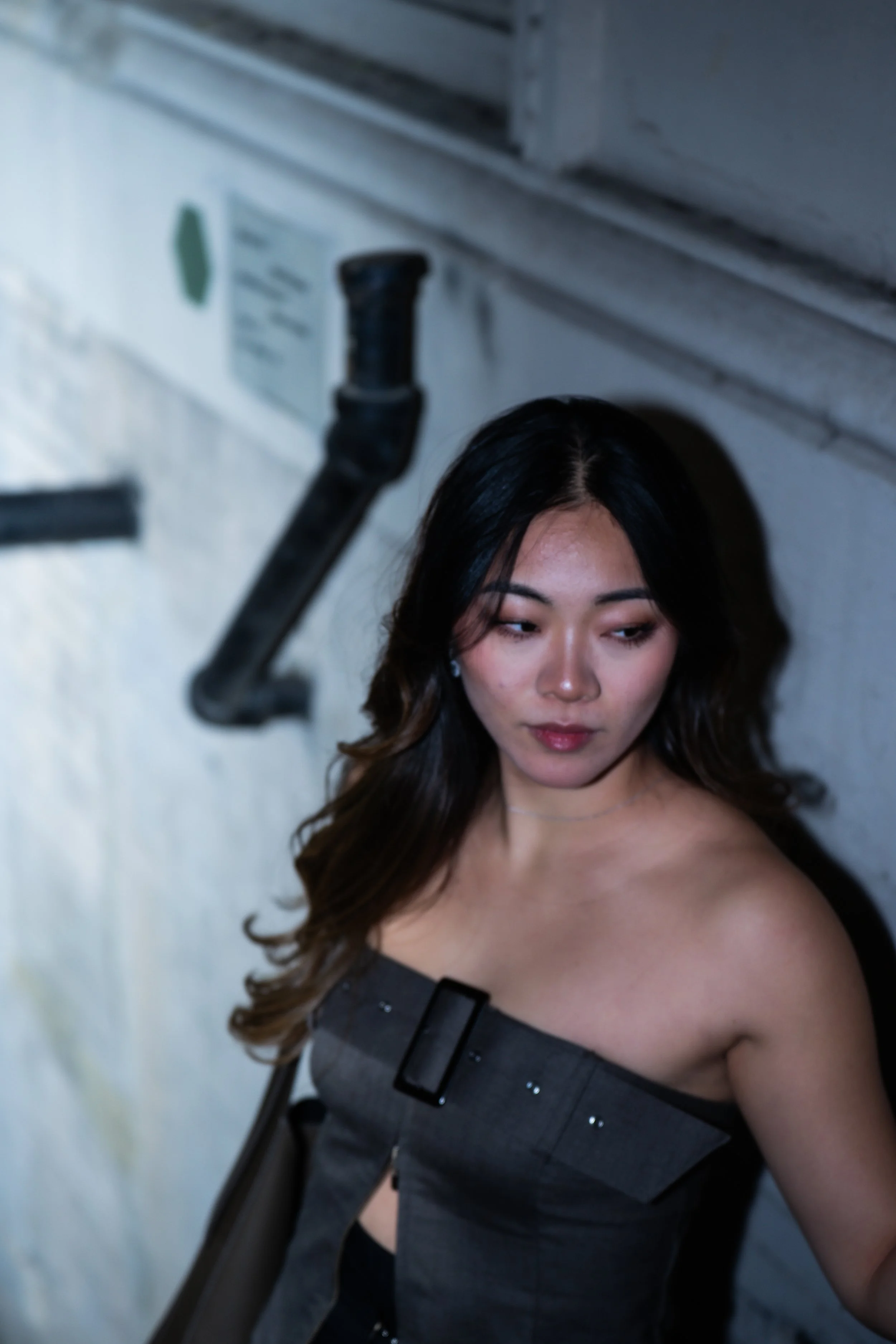A young woman with long dark hair, wearing a strapless dark dress, posing against a white brick wall with a black pipe overhead.