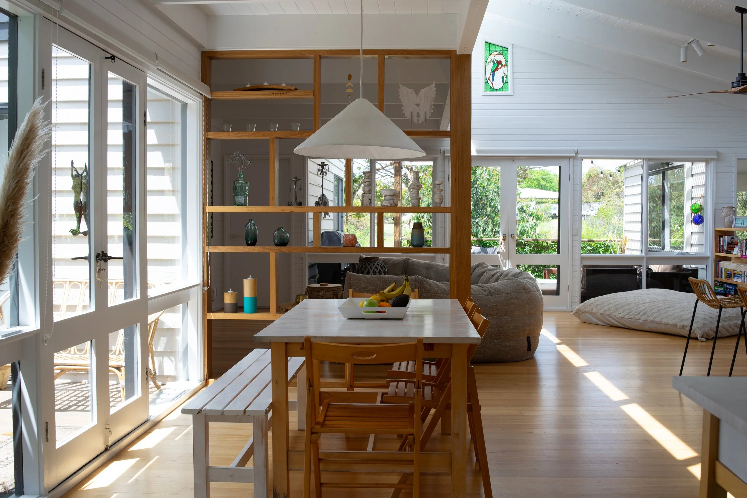 Bright, open living room with large windows, wooden flooring, a dining table with chairs, a sofa, and a dog bed, decorated with artwork and plants.
