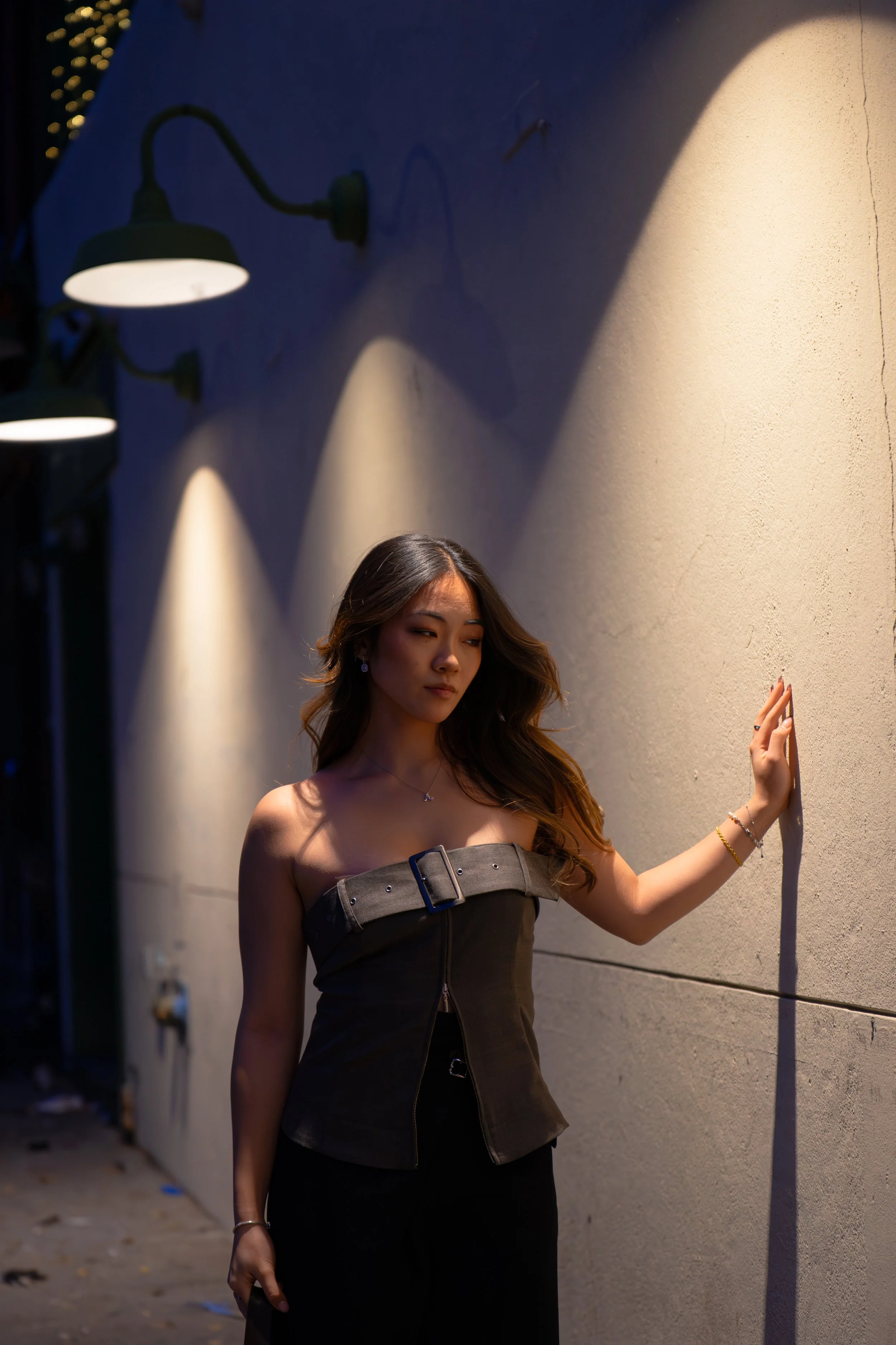 A young woman standing outside at night, leaning against a beige wall with her hand, illuminated by wall-mounted lights.
