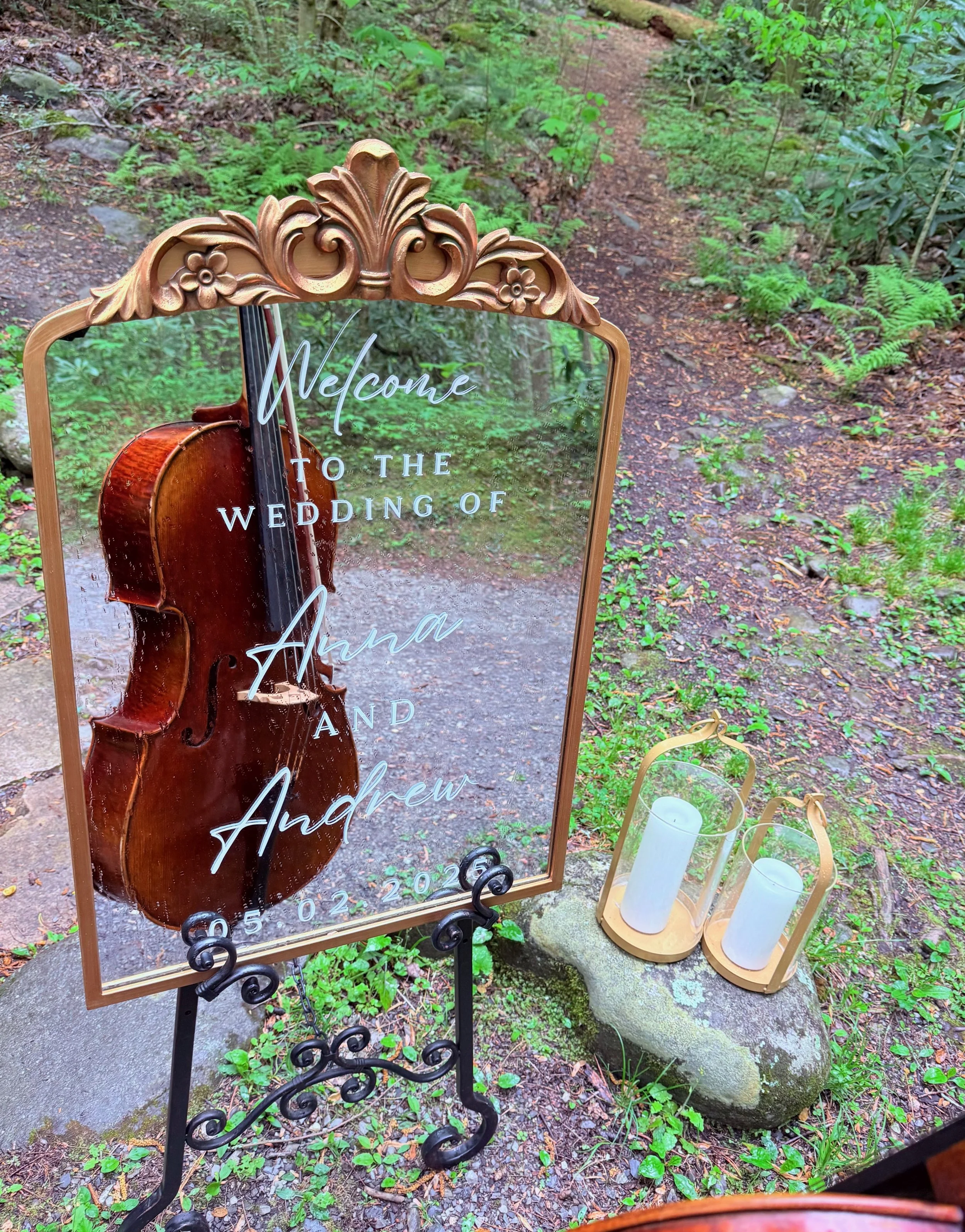 A wedding welcome sign with a mirror design and a wooden ornate frame, reflecting a violin, placed outdoors on a trail surrounded by green foliage. The sign reads, 'Welcome to the wedding of Alana and Andrew 05.02.2020', with two white candles in lan