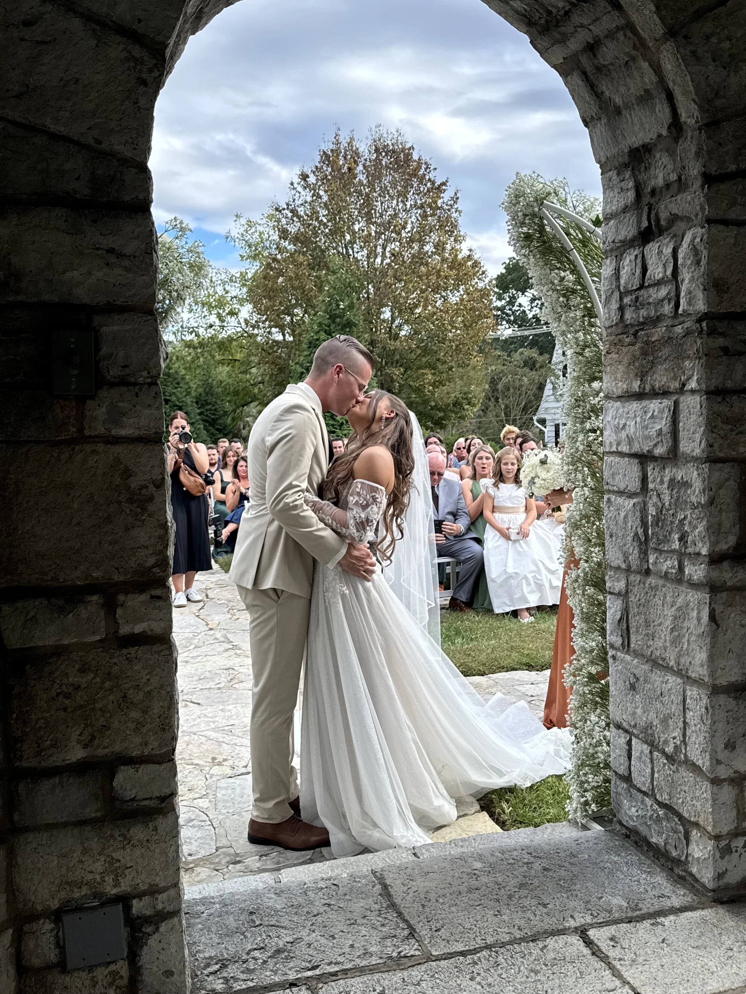 A couple shares a kiss during their wedding ceremony, viewed through a stone archway. The bride wears a white gown and veil, while the groom is in a light-colored suit. Guests are seated, watching the moment, with some taking photos, amid trees and a cloudy sky.