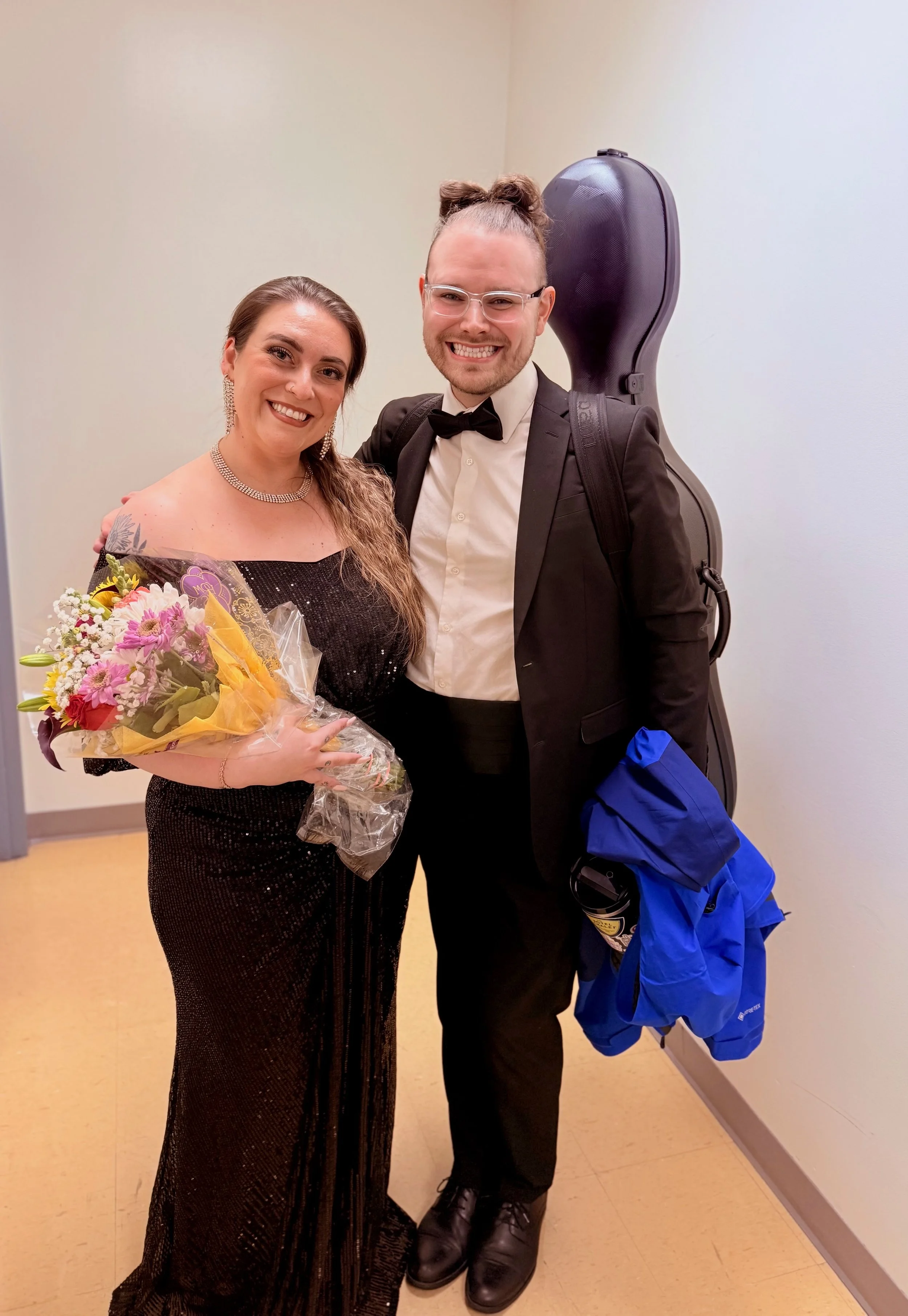A woman in a black, sequined gown holding a bouquet of flowers, standing next to a man in a tuxedo with a guitar case on his back, both smiling.