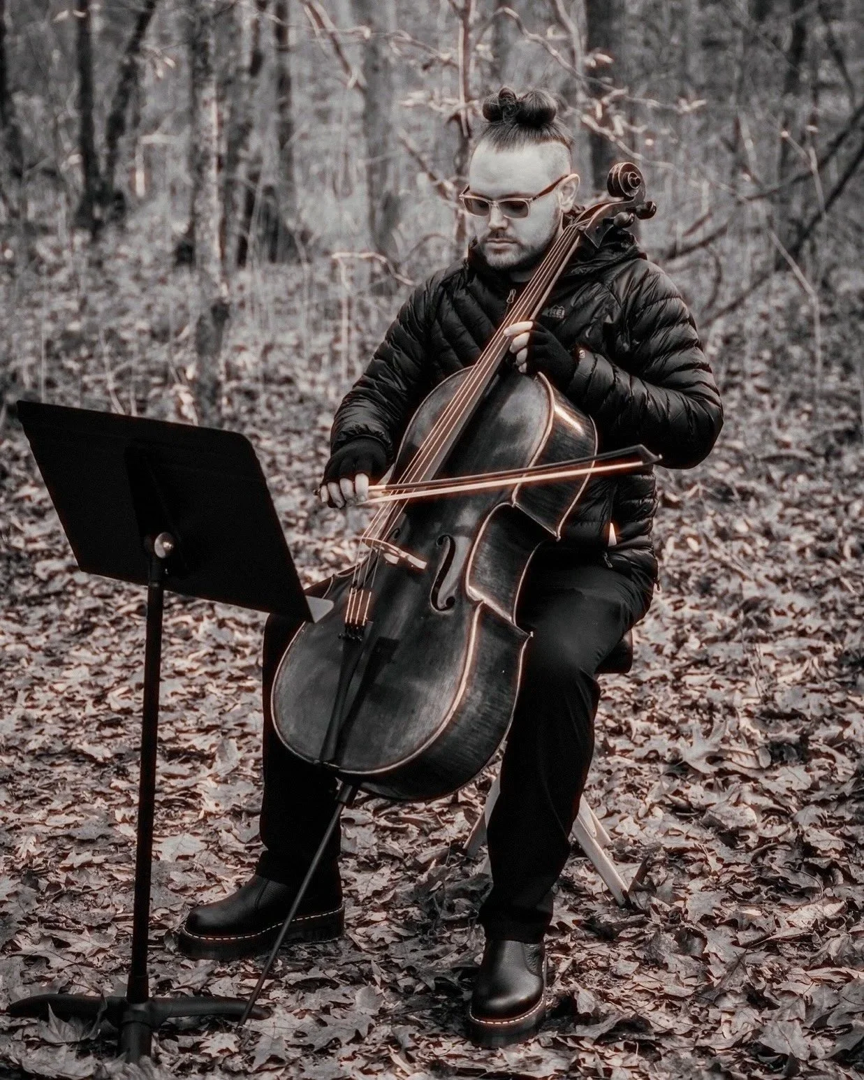 A man wearing glasses and a black jacket is sitting outdoors on a fallen leaf-covered ground, playing a cello with a music stand nearby, surrounded by trees in a forest setting.