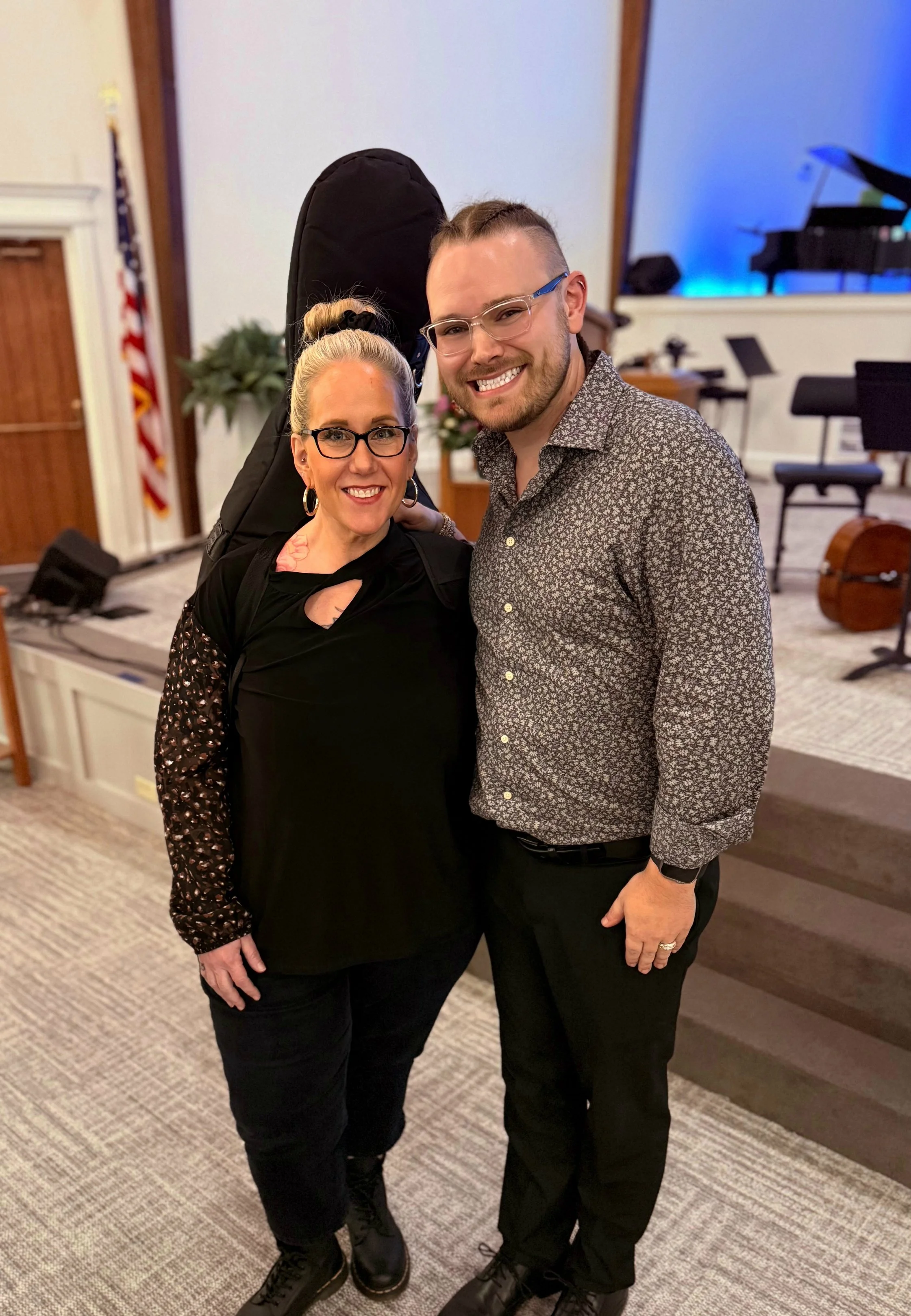 A woman and a man smiling, standing close together in a room with musical instruments and a stage in the background. The woman has blonde hair in a bun, wears glasses, a black top with patterned sleeves, and black pants. The man has short hair, glass