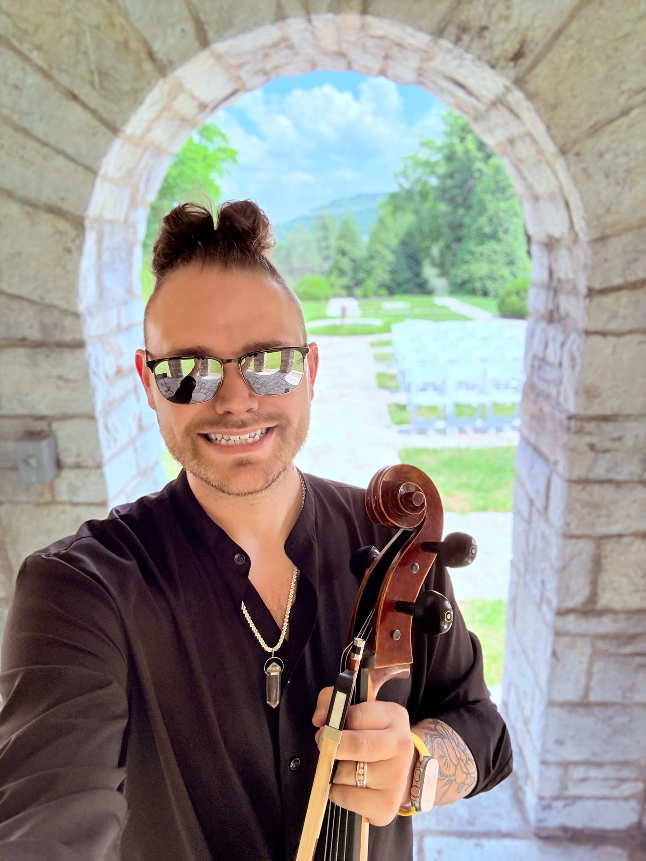 A man with sunglasses and a bun hairstyle takes a selfie while holding a violin, standing in front of a stone archway with a scenic outdoor view of greenery and blue sky in the background.