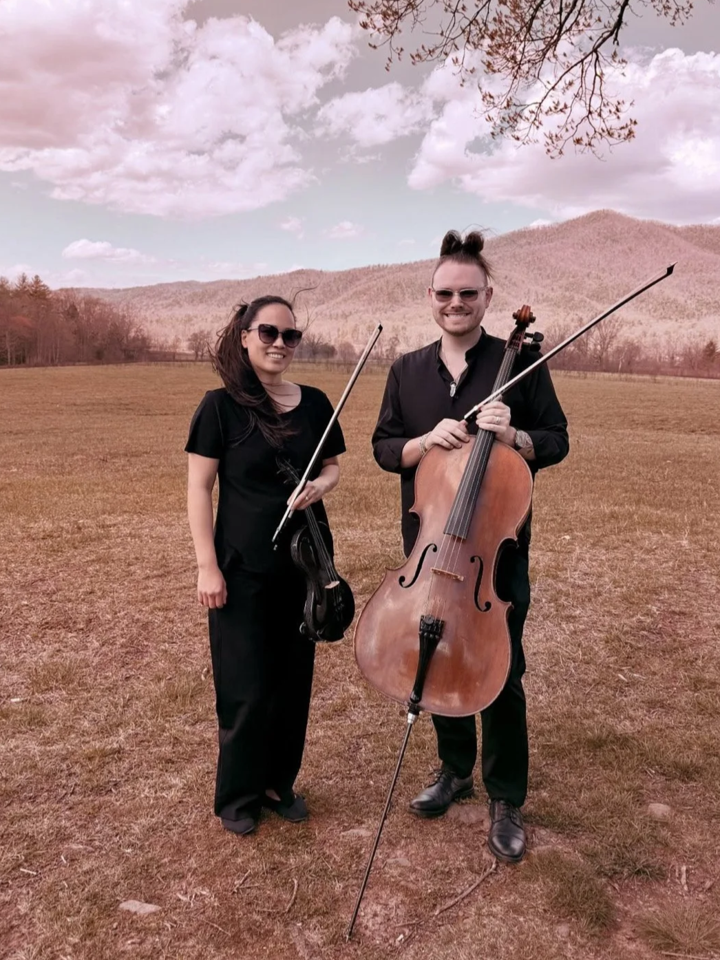 Two musicians, a woman and a man, standing outdoors in an open field with mountains in the background. They are holding violins and a cello and are smiling at the camera, wearing sunglasses and black clothing.