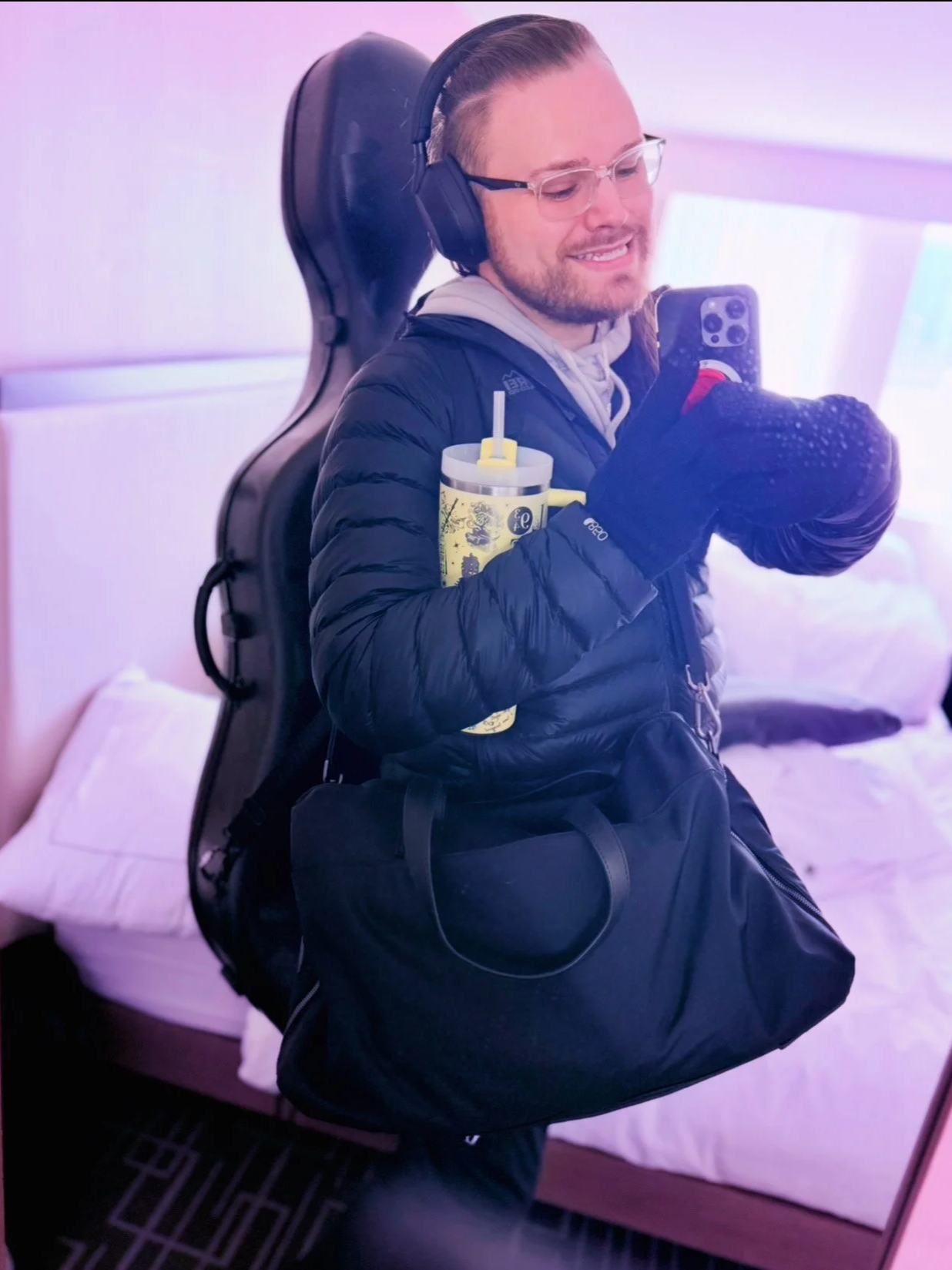 A young man with glasses and a beard taking a selfie in a hotel room mirror. He is wearing a black puffy jacket and black gloves, holding a sports water bottle with a straw, a black shoulder bag, and is leaning against a suitcase. He is smiling and w