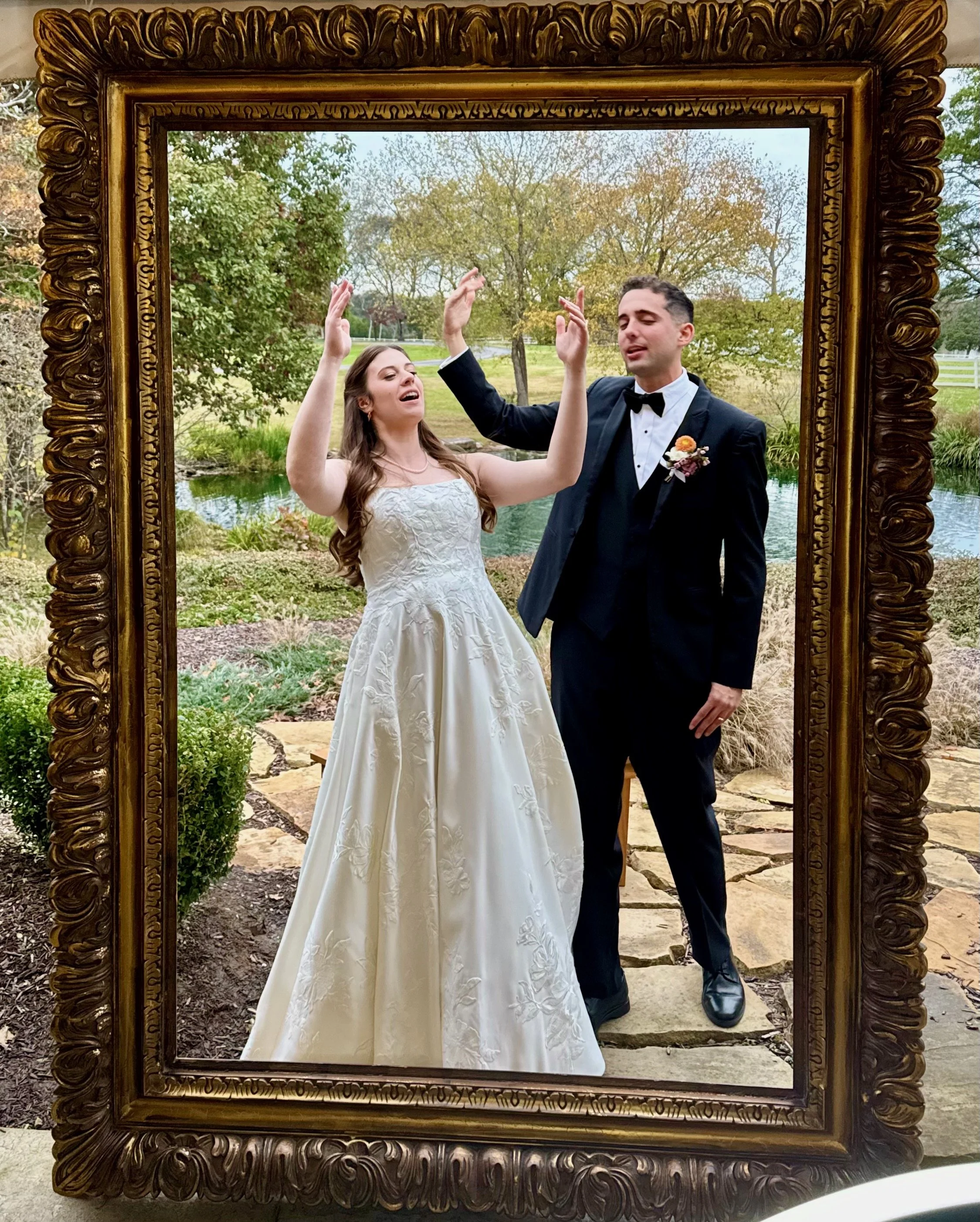 A bride and groom inside a decorative gold frame, celebrating their wedding outdoors by a pond with trees and greenery in the background.