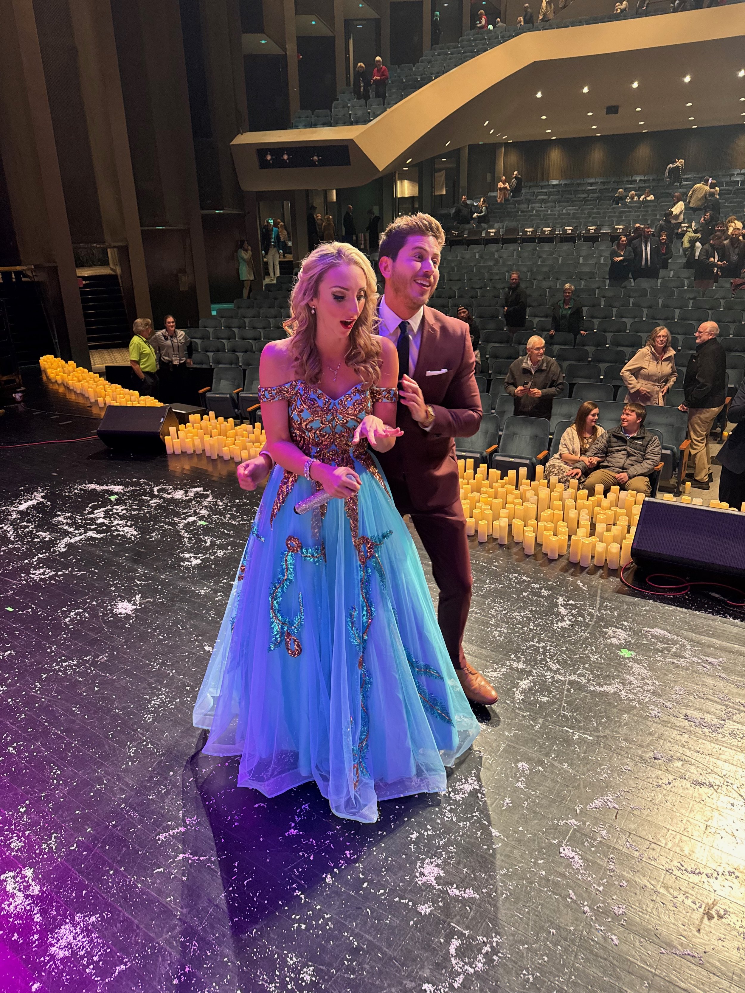 A young woman in a colorful, sparkly ball gown and a young man in a suit are on stage, moments after their engagement live on stage! The background shows empty auditorium seats and a few spectators.