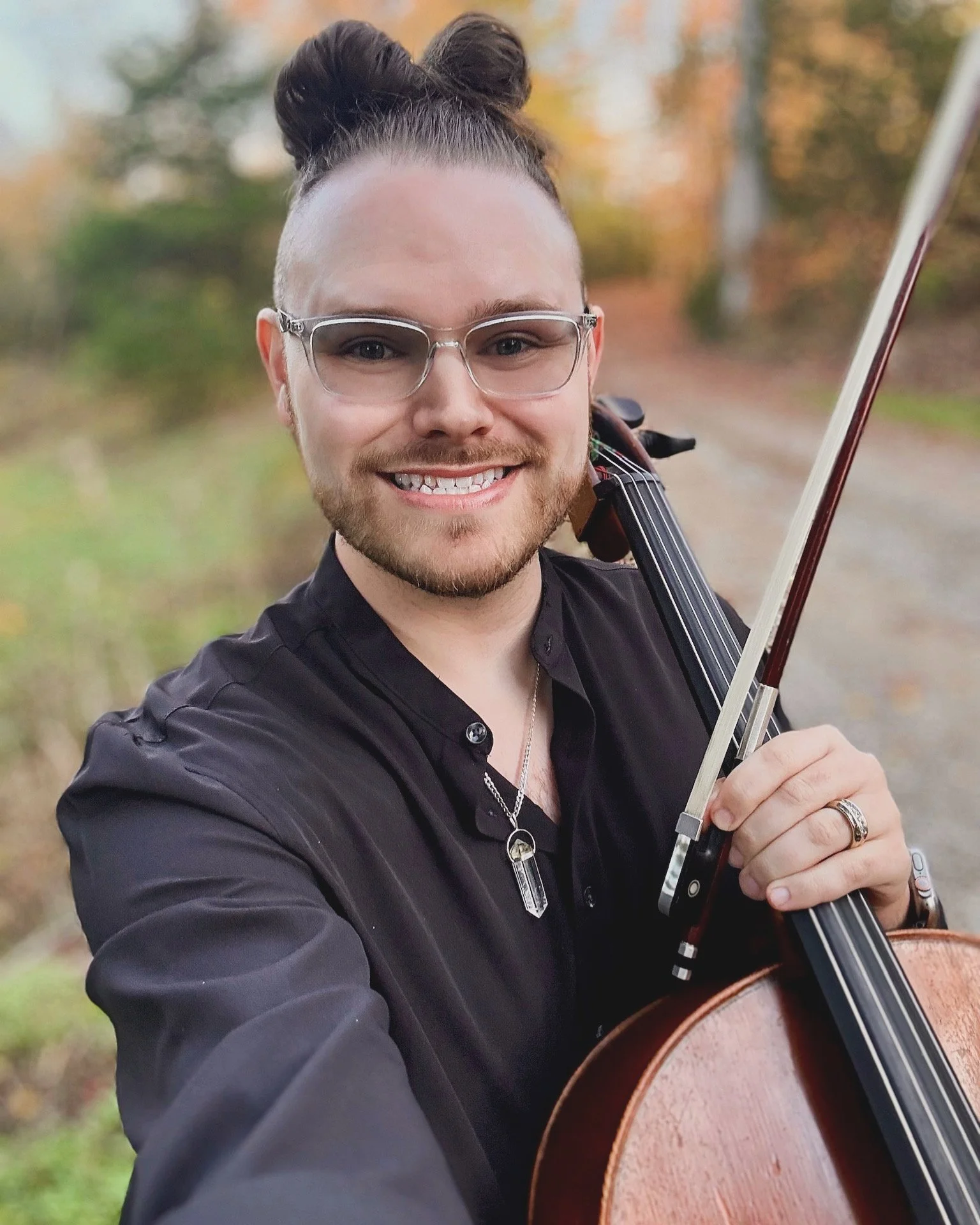 A smiling person with glasses, earrings, and a necklace, holding a cello outdoors during autumn, with trees and a dirt path in the background.