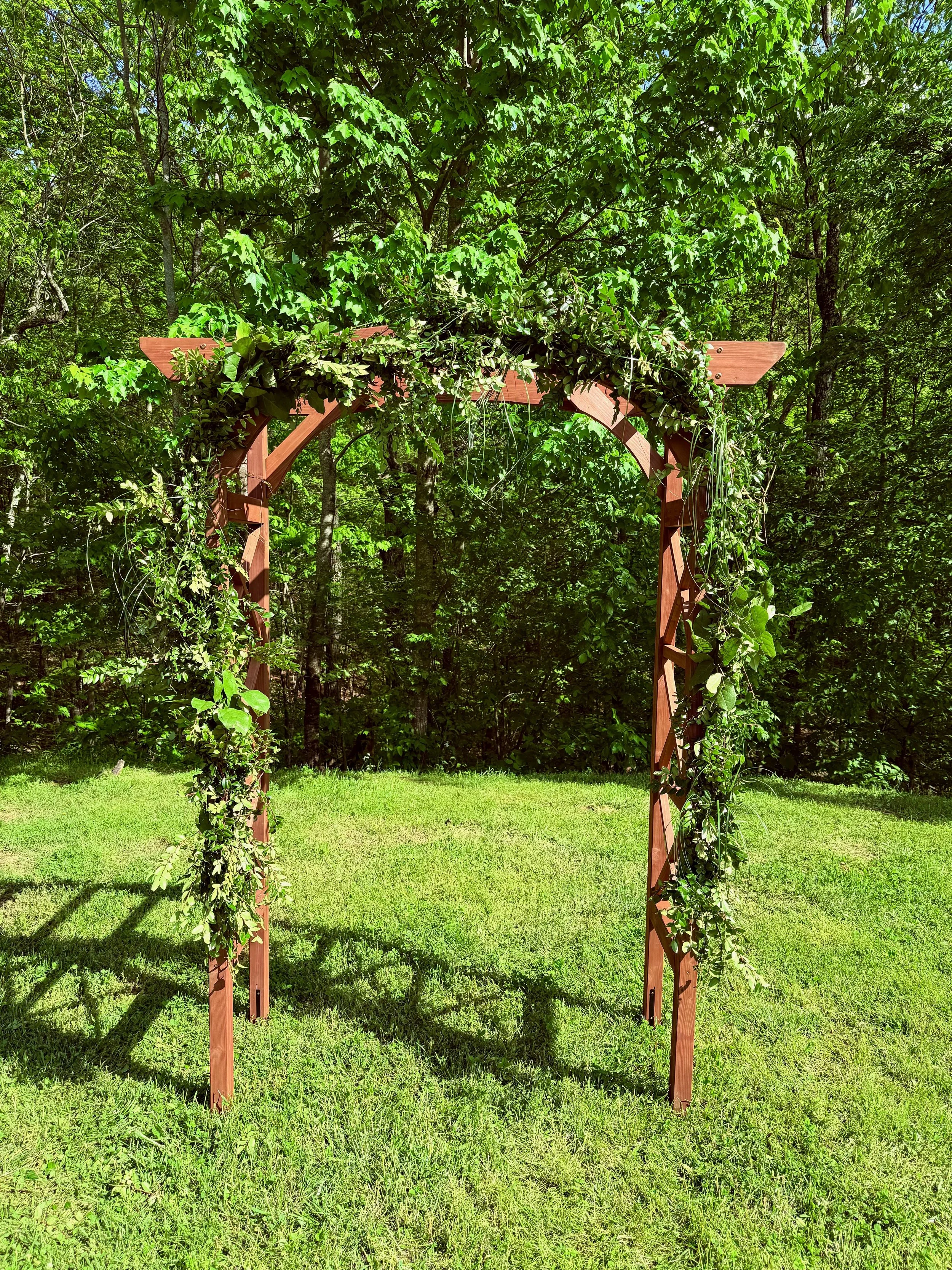 Wooden arch decorated with green leaves and vines set on a grassy area in a garden or park with trees in the background.