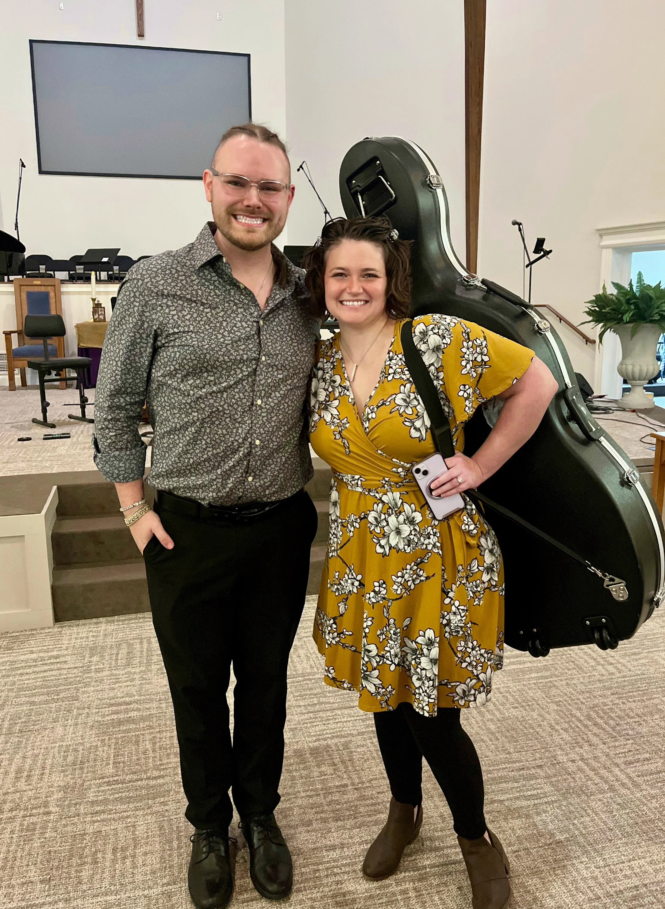 A man and a woman stand together smiling inside a church, with a musical instrument case behind them. The man has glasses and a patterned shirt, the woman is wearing a yellow floral dress and holding a smartphone.