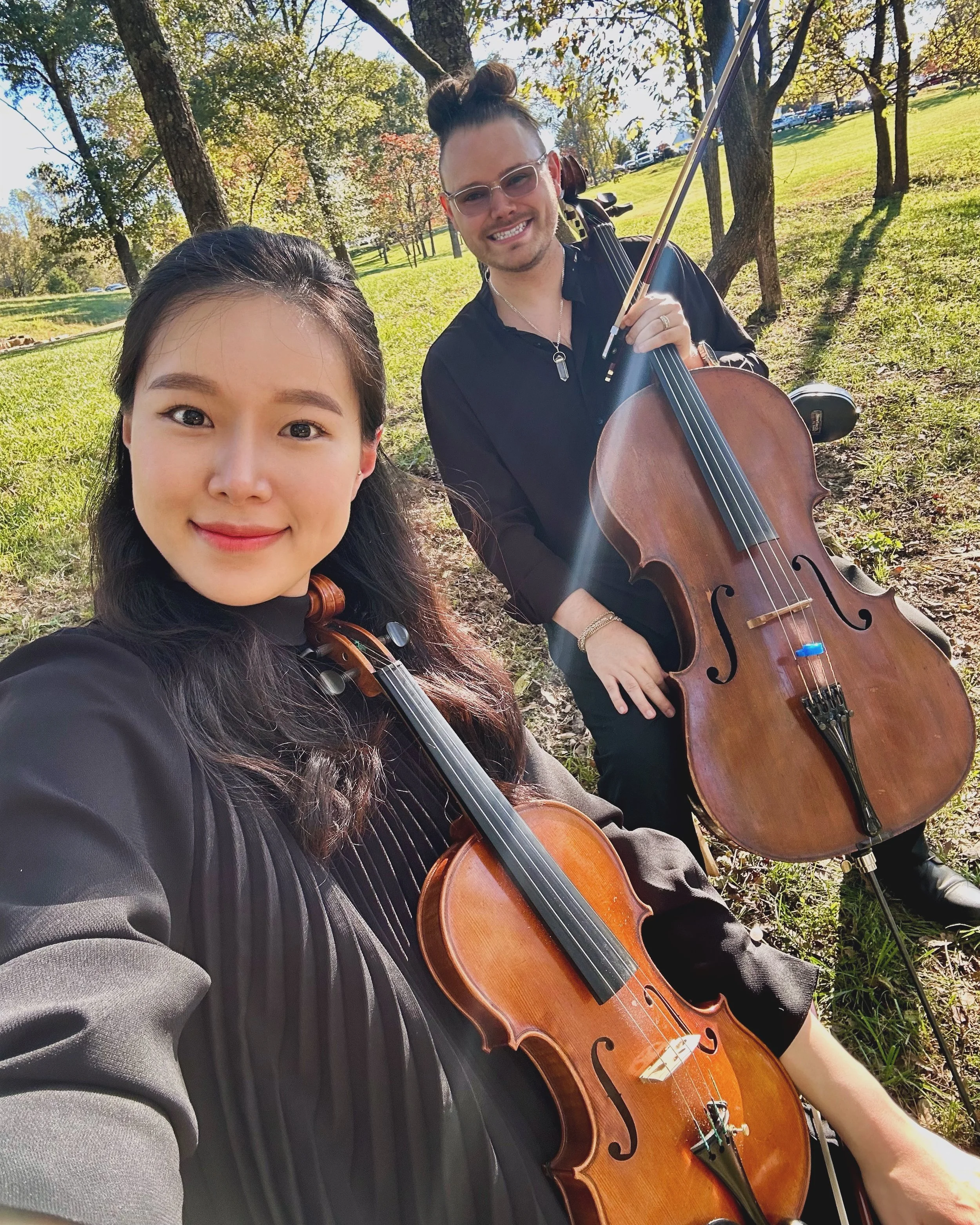 Two musicians, a woman with a violin and a man with a cello, sitting outdoors on grass with trees and sunshine in the background.