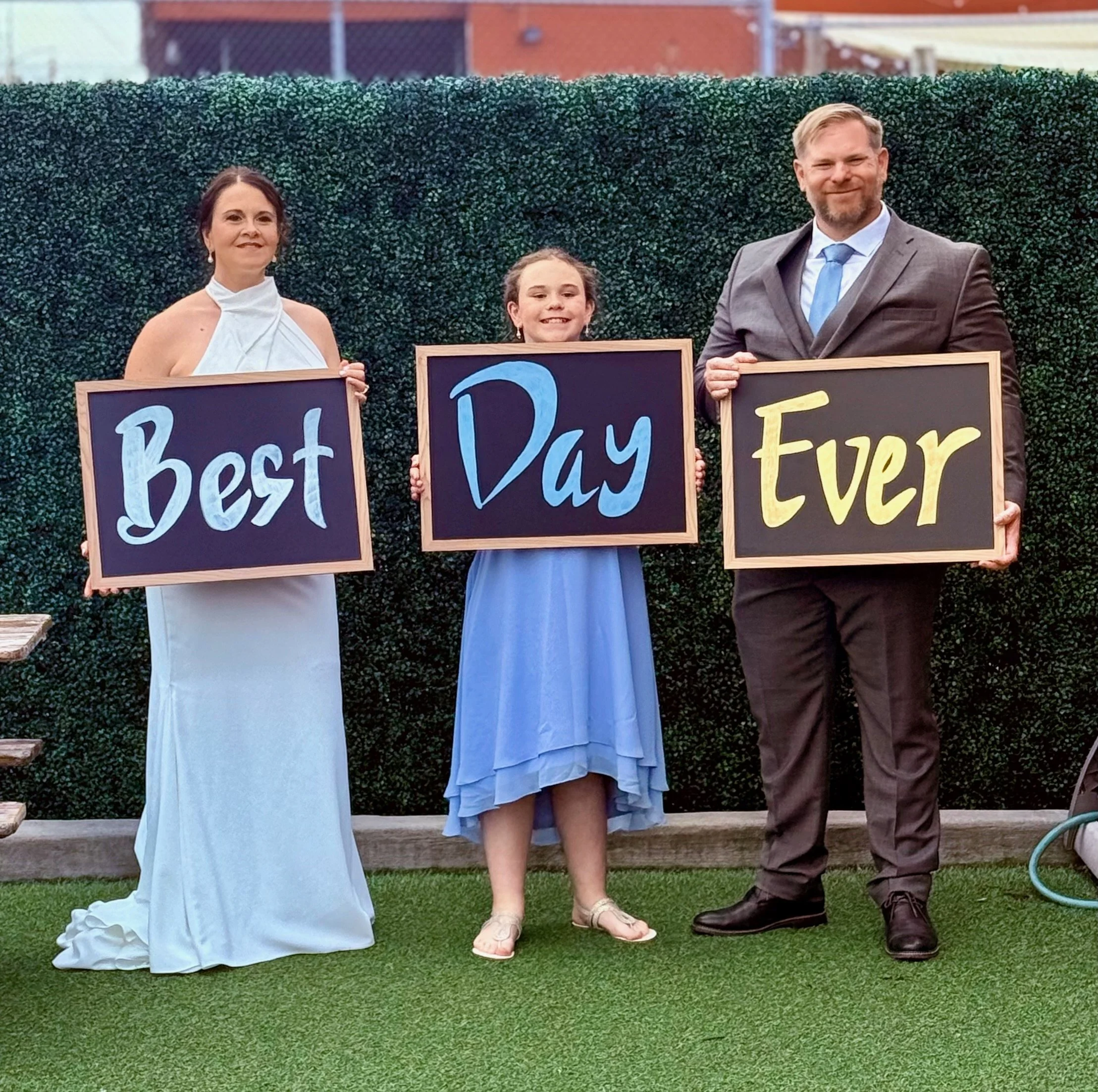 Three people standing outdoors in front of a hedge, holding signs that spell out 'Best Day Ever'. A woman in a white dress on the left, a girl in a blue dress in the middle, and a man in a suit on the right.