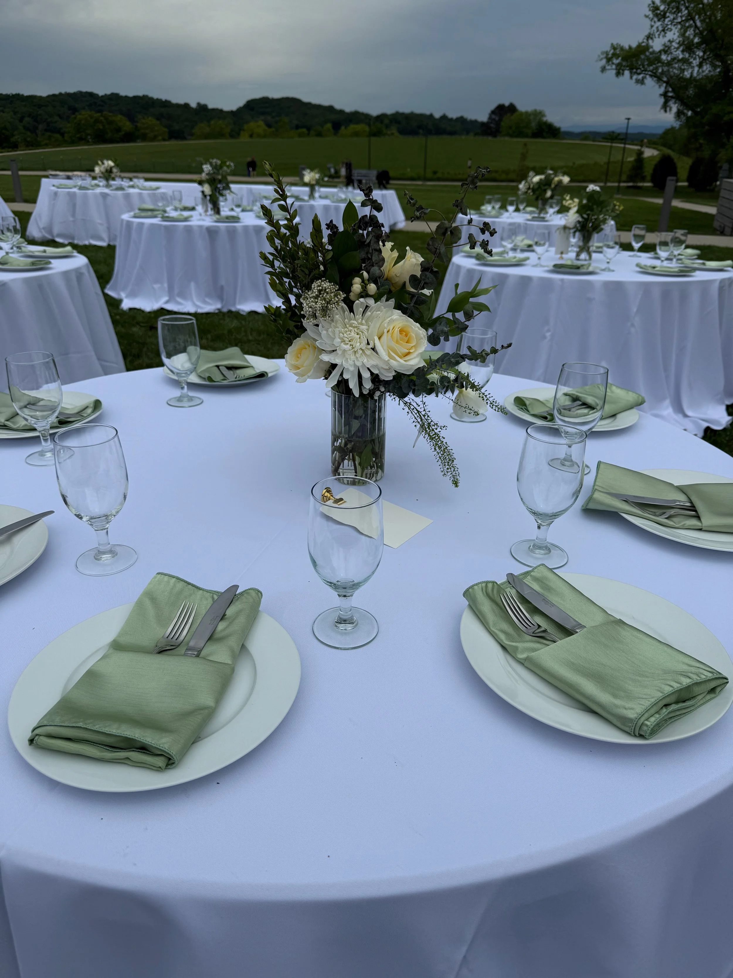 Outdoor wedding reception tables with white tablecloths, green napkins, glassware, and floral centerpieces featuring white roses and other flowers, set on a grassy area with rolling hills in the background.