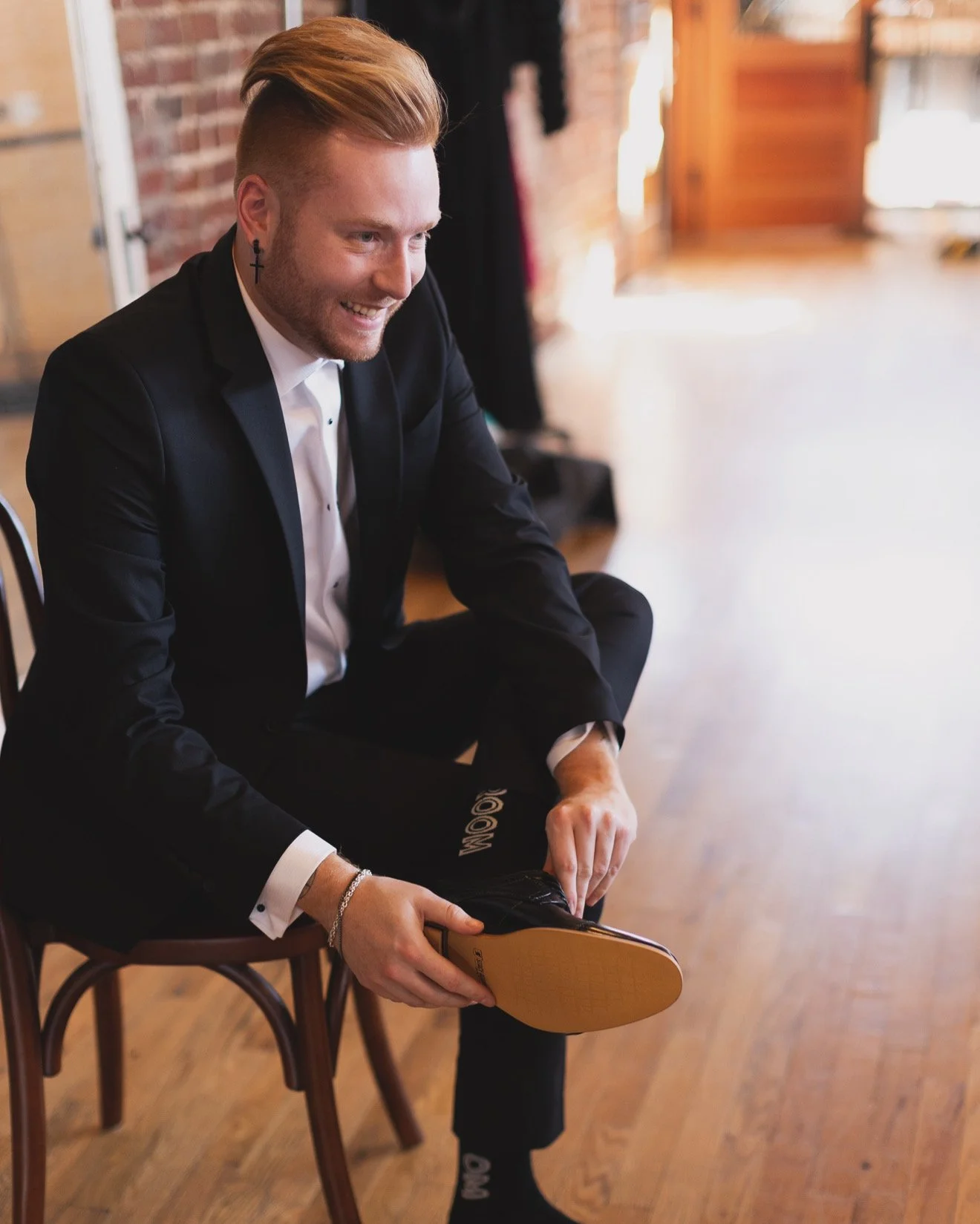 A young man with styled blonde hair, wearing a black suit and white shirt, is sitting on a wooden chair, smiling while putting on a black dress shoe.