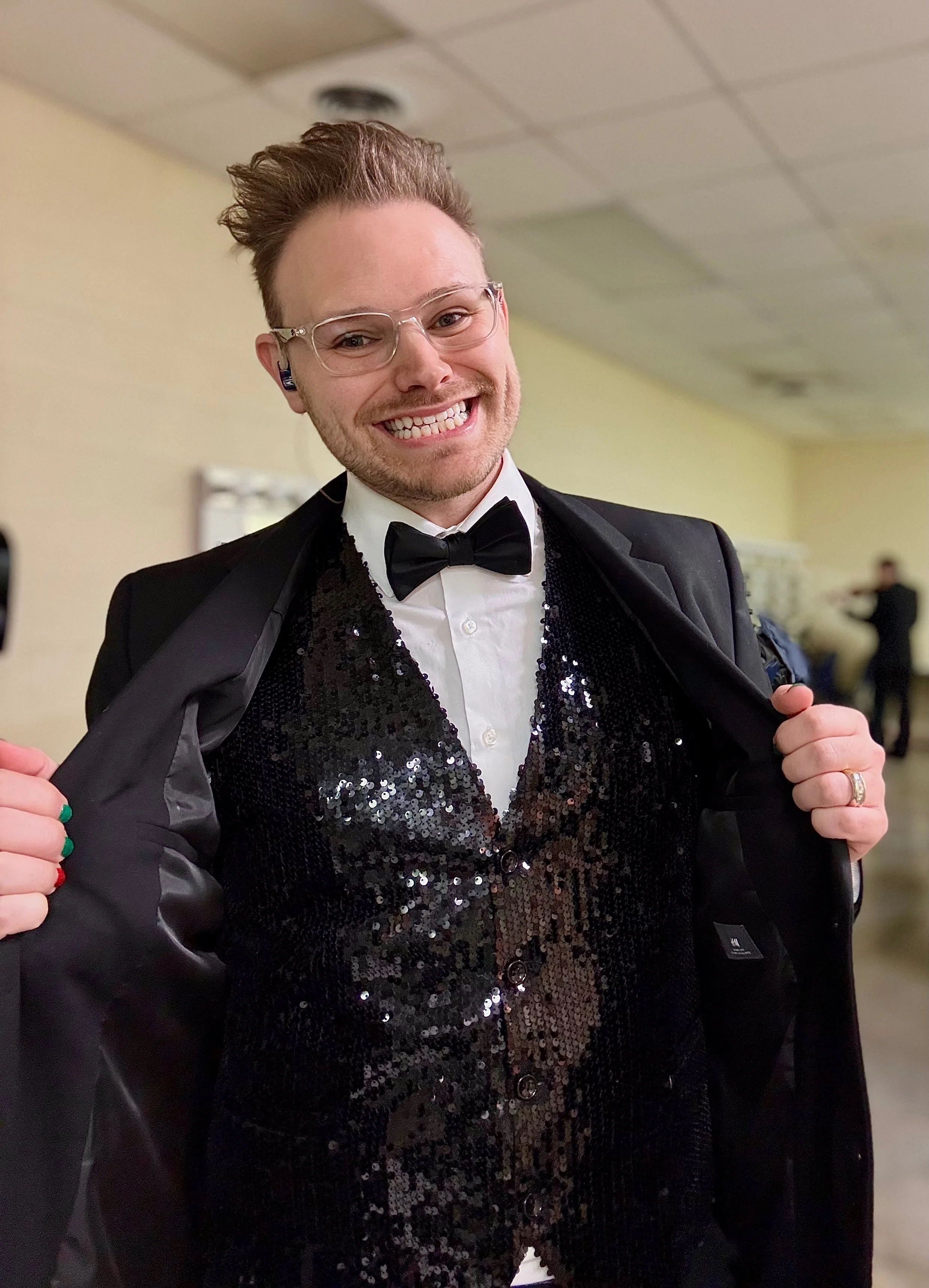A man in formal wear, including a black tuxedo with a sequined vest, white dress shirt, and black bow tie, smiling and holding open his jacket. He has glasses, styled hair, and is standing indoors at an event.