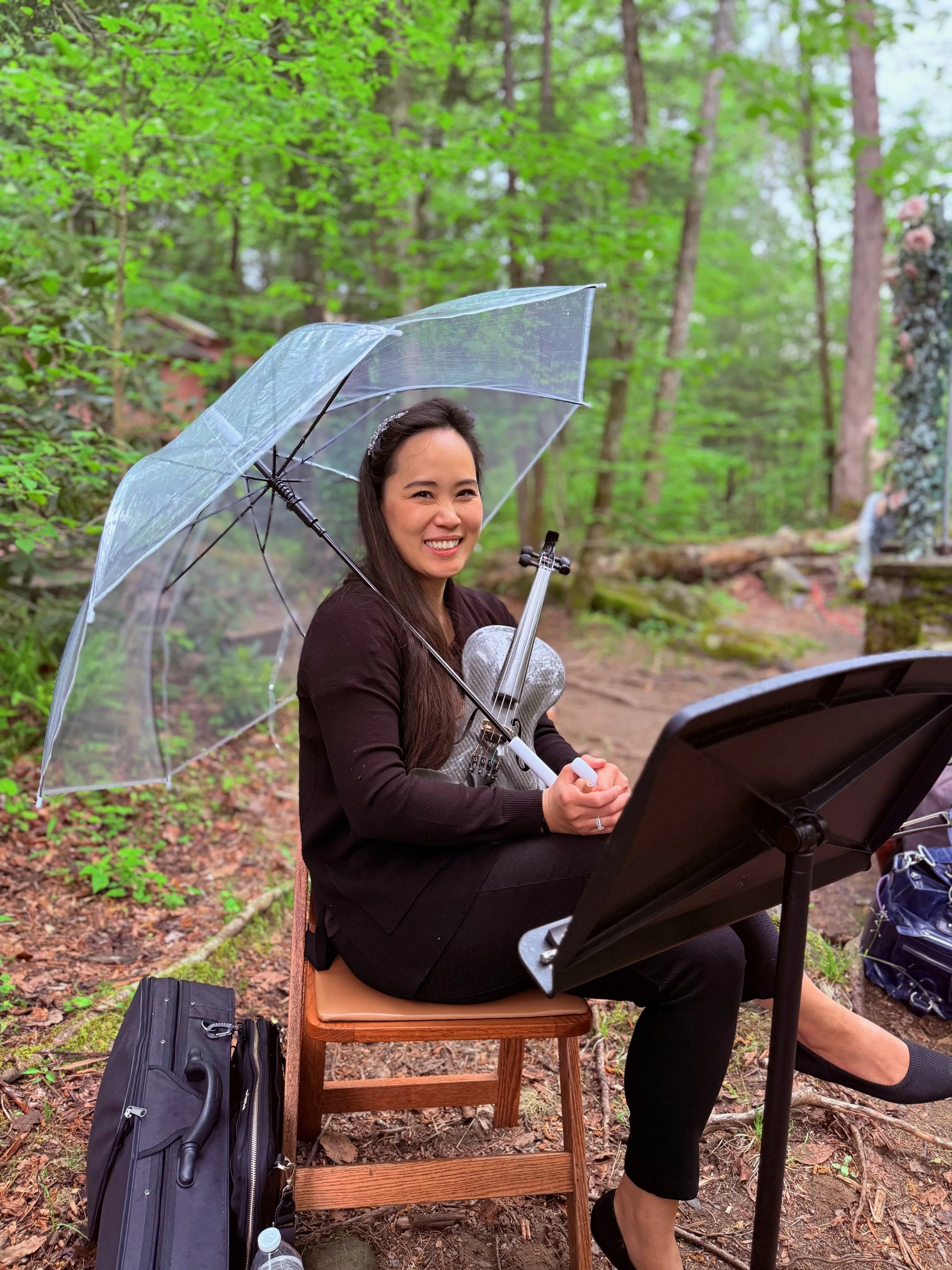 A woman sitting outdoors in a forested area, smiling while holding a violin under a clear umbrella, with music stand and sheet music in front of her.