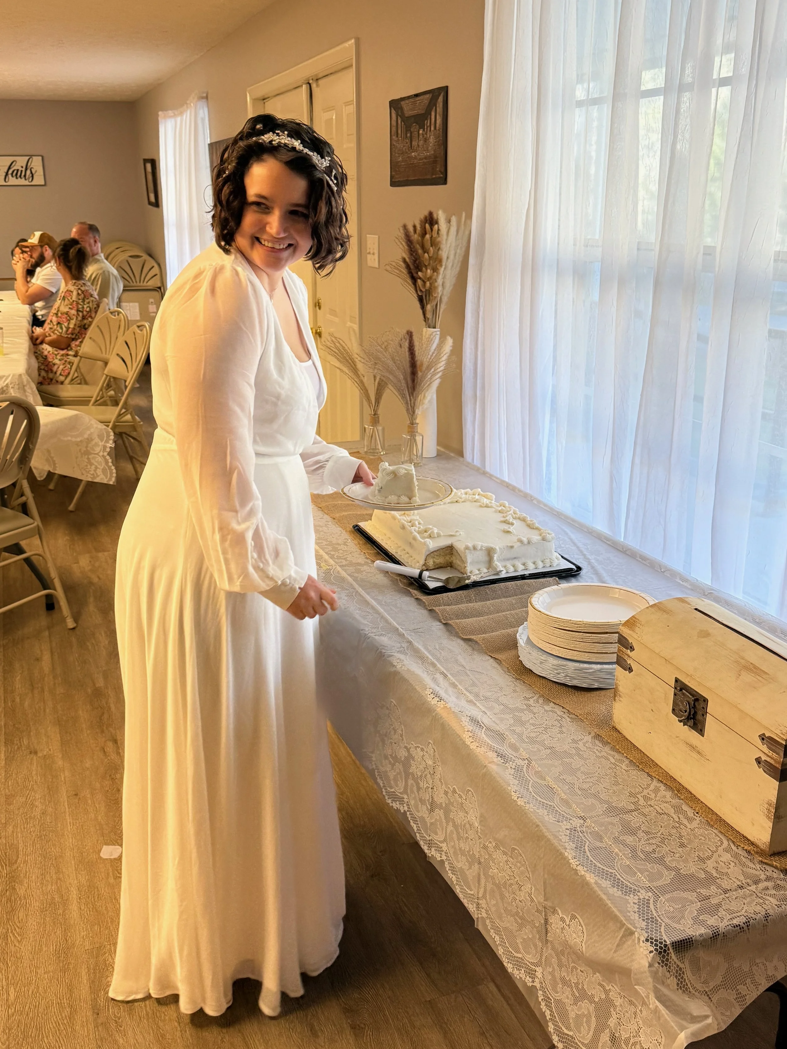 A woman in a white dress is cutting a cake at a celebration, with a happy expression on her face. There are plates and a wooden box on the table.