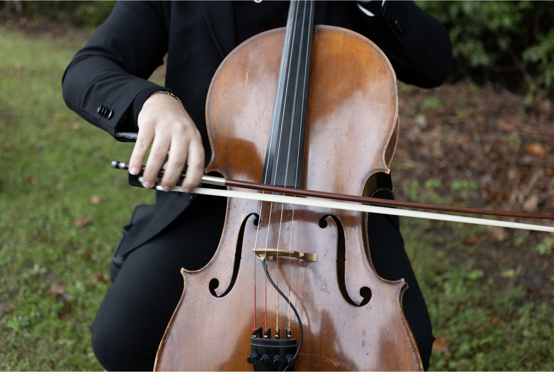 Close-up of a person playing a cello outdoors, wearing a black suit, with a blurred green and brown background.