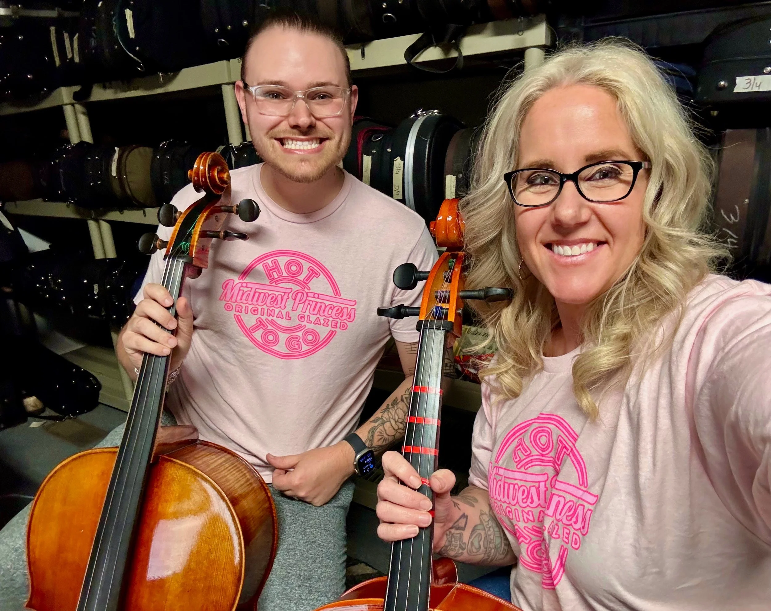 Two smiling people, a man and a woman, holding violins in a music store. They are wearing matching pink T-shirts that say 'Midwest Princess Original Glazed' with a circular logo. The background shows shelves filled with musical instrument cases.