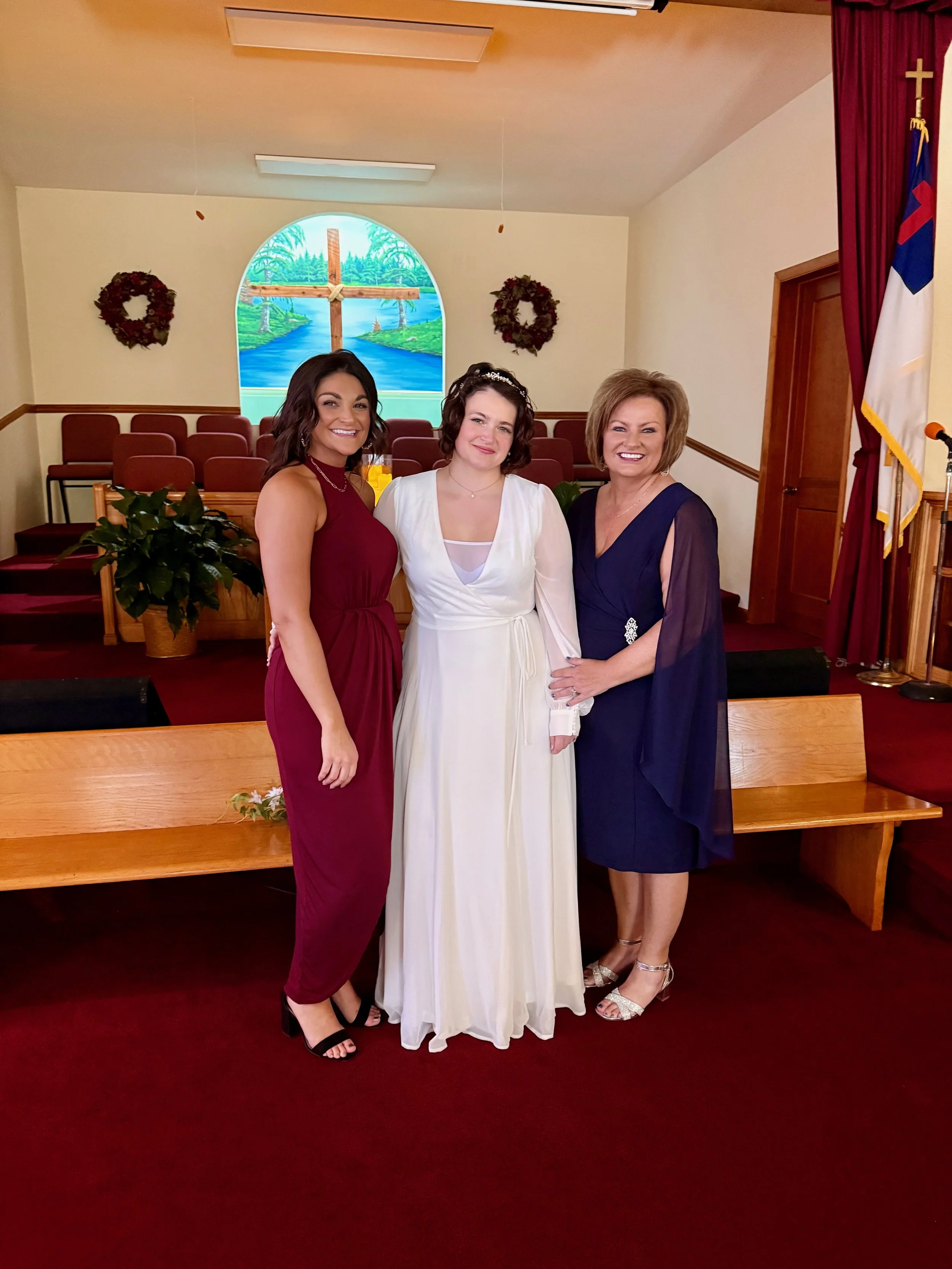Three women standing together in a church, with a stained glass window depicting a cross and scenic background behind them. The woman in the center wears a white dress, the woman on the left wears a maroon dress, and the woman on the right wears a na