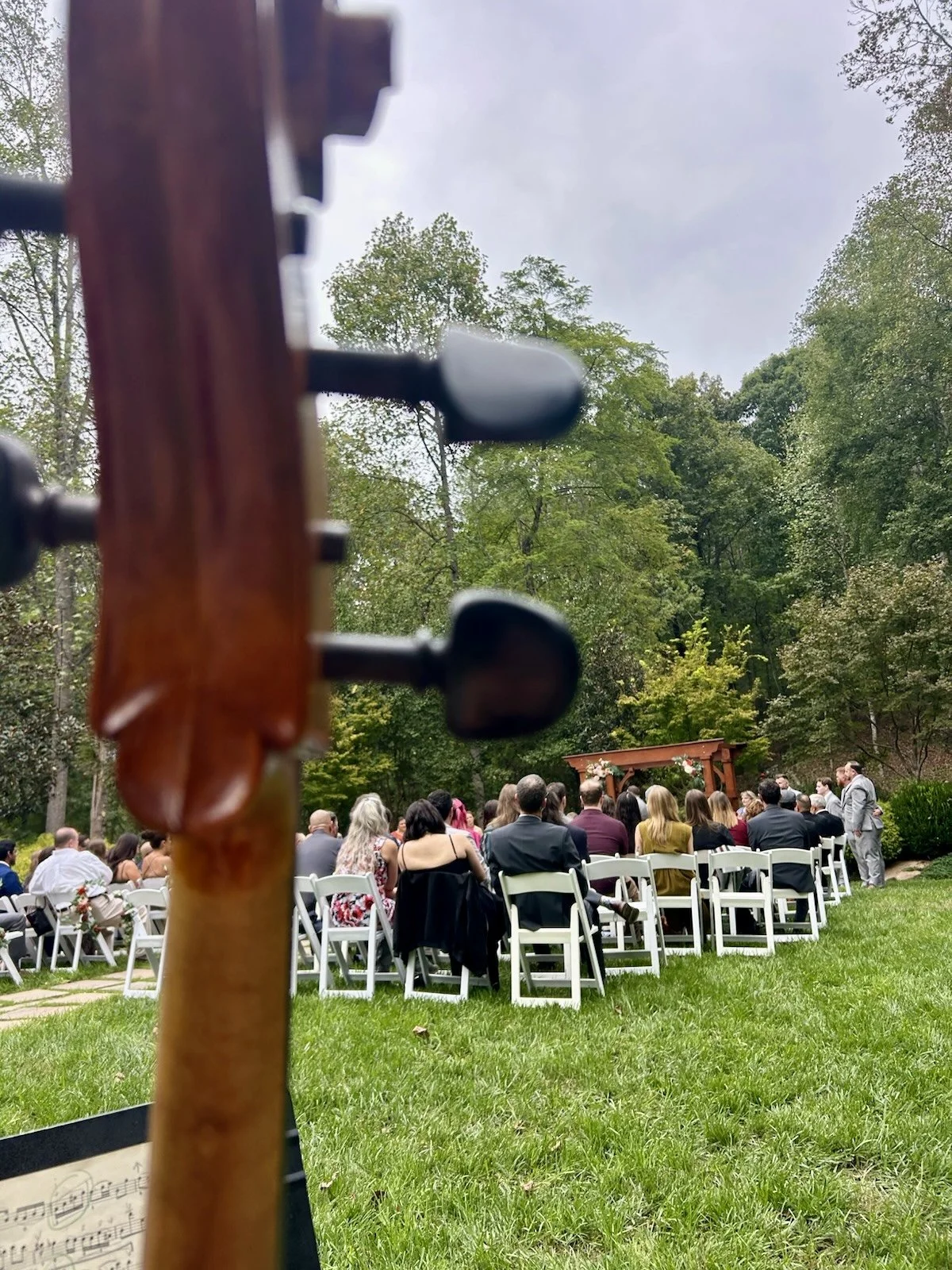 A group of people gathered outdoors for a wedding ceremony, seated under a canopy in a wooded area with green grass and trees, viewed from behind a guitar.