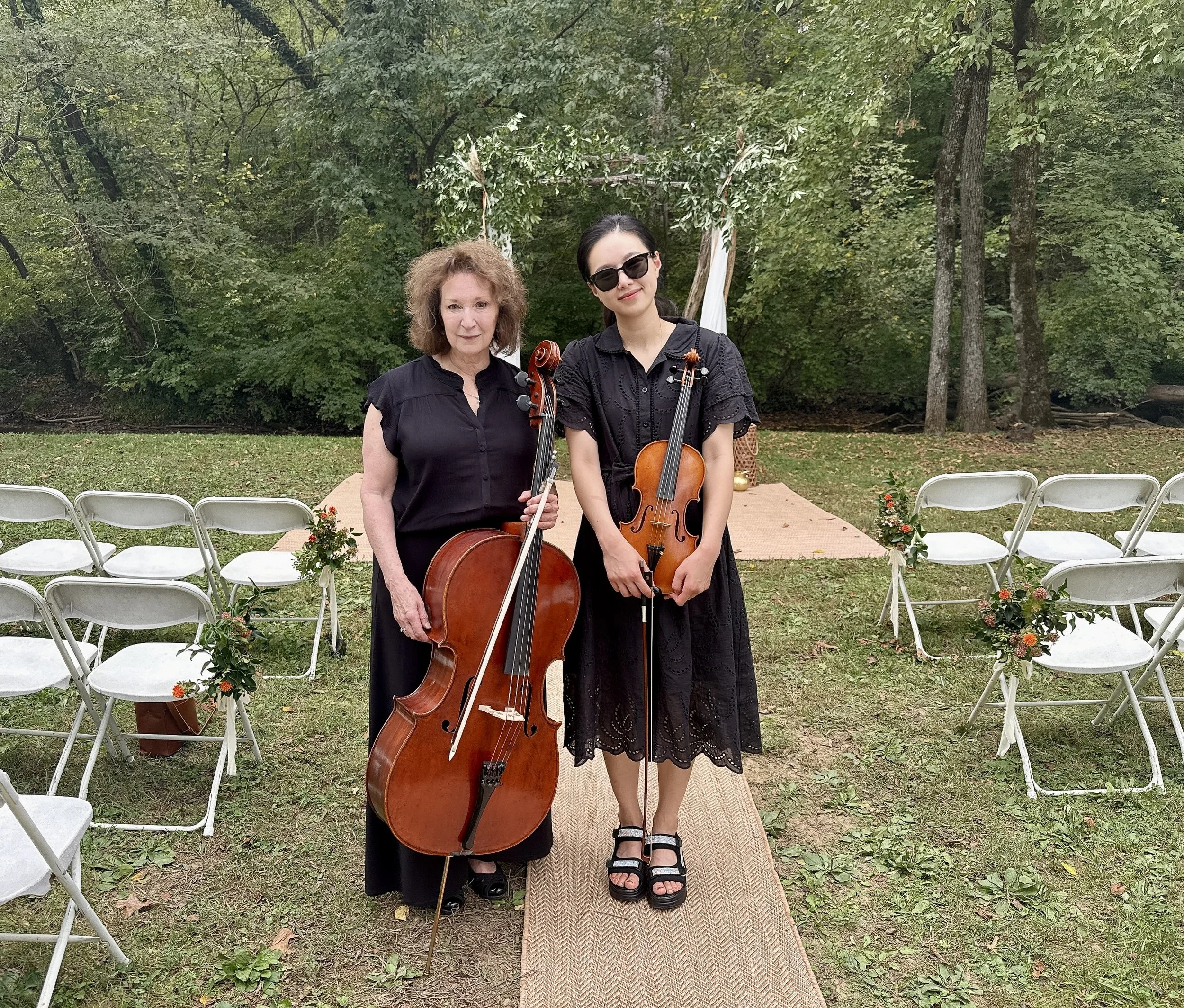 Two women holding string instruments, standing on an outdoor aisle decorated with flowers, with chairs arranged on both sides and a wooded area in the background.
