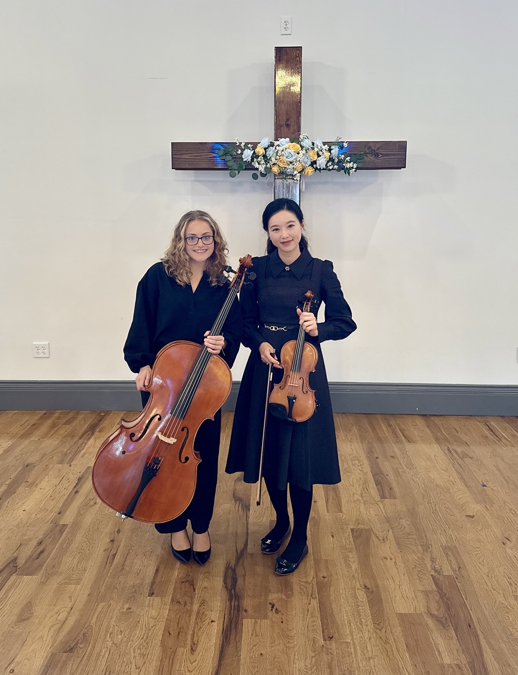 Two women holding string instruments, standing in front of a wooden cross with a floral arrangement, on a wooden floor.