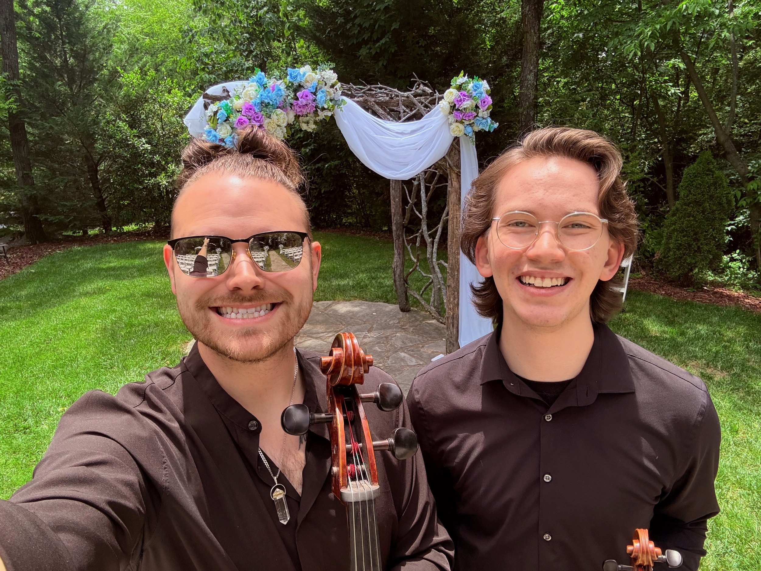 Two smiling young men stand outdoors in front of a decorated arch with white fabric and colorful flowers, one holding a violin and both wearing black shirts.