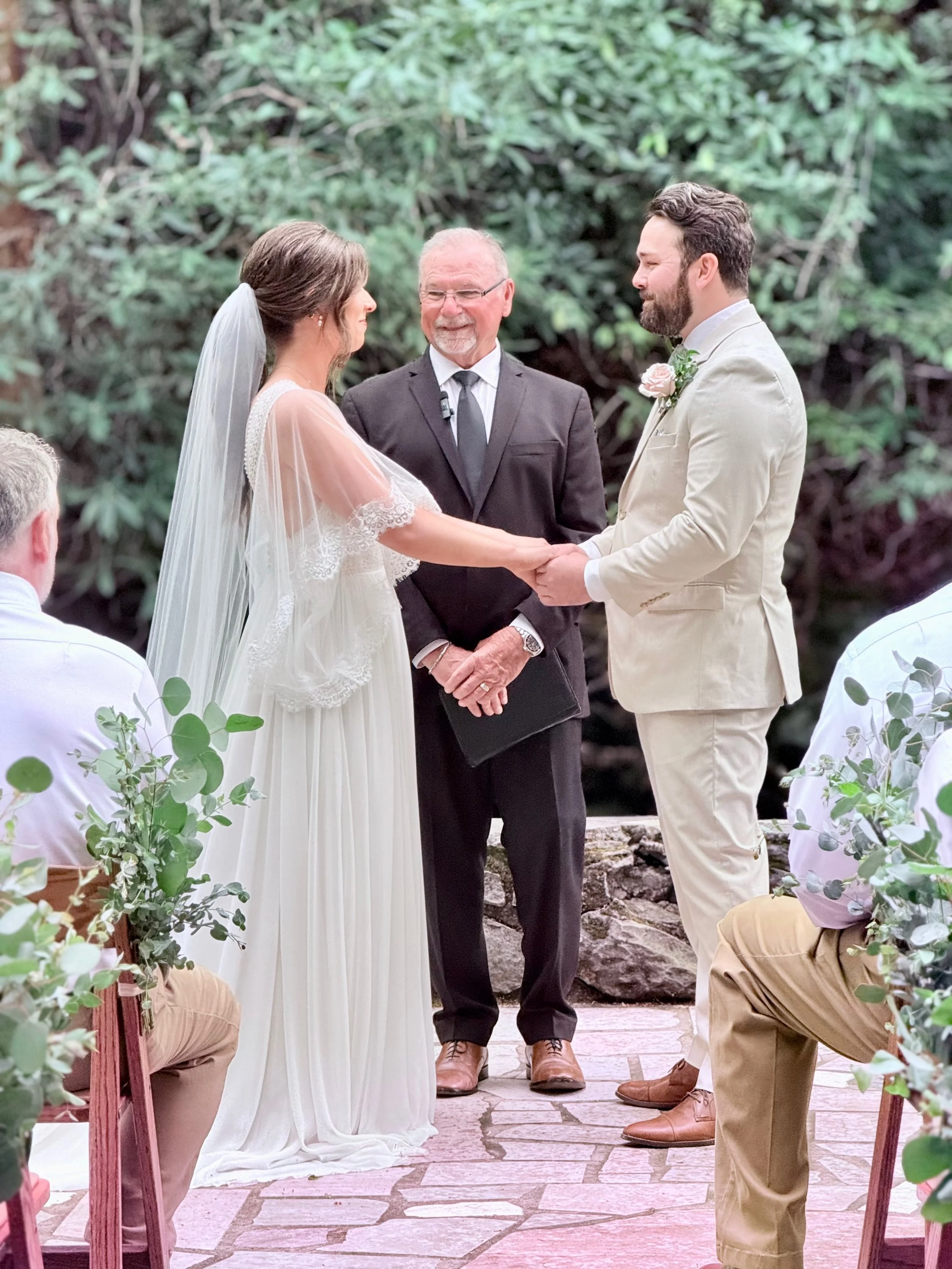 A bride and groom holding hands during a wedding ceremony outdoors, with an officiant officiating, surrounded by seated guests and greenery.