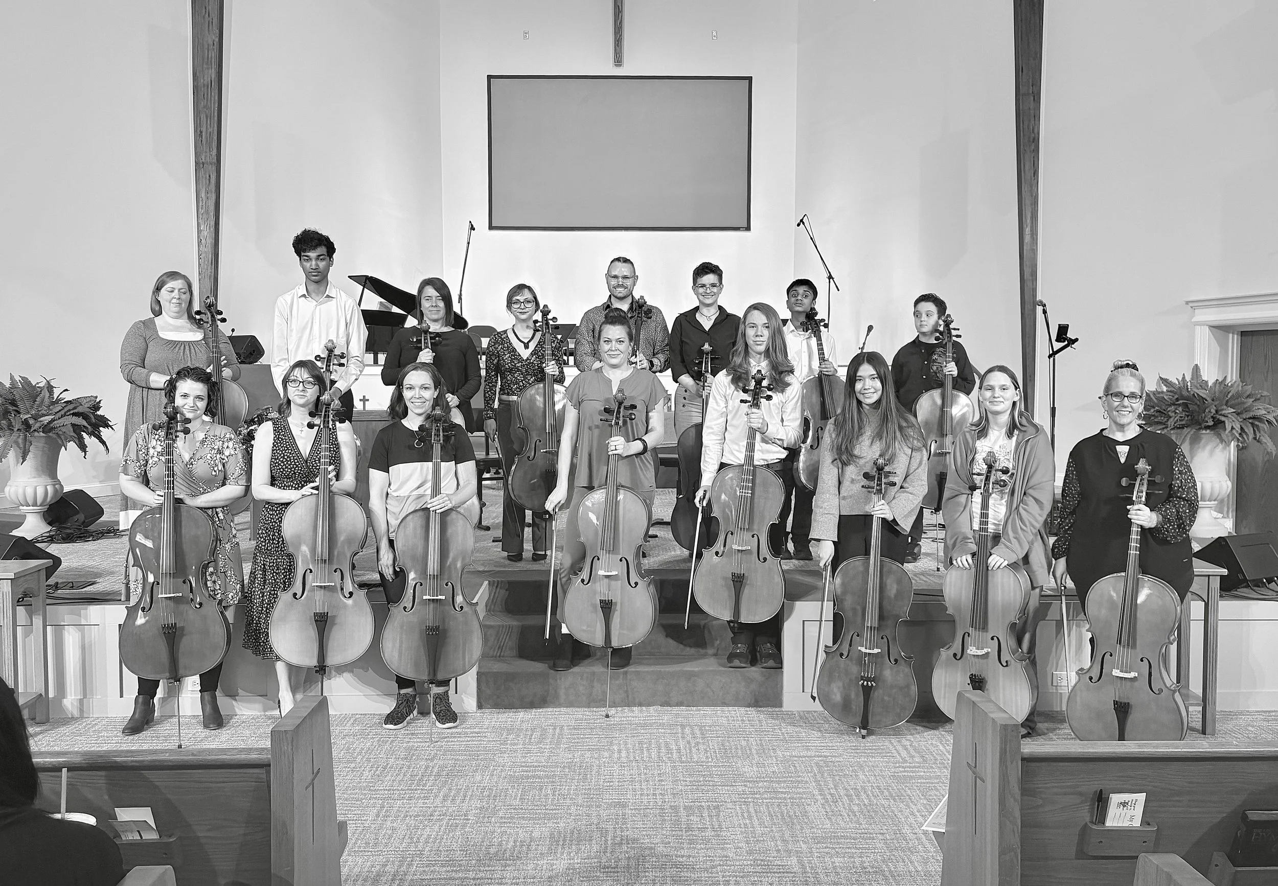 Black and white photo of a group of musicians holding string instruments, standing on a stage inside a church or auditorium. They are arranged in two rows, with some seated and others standing.