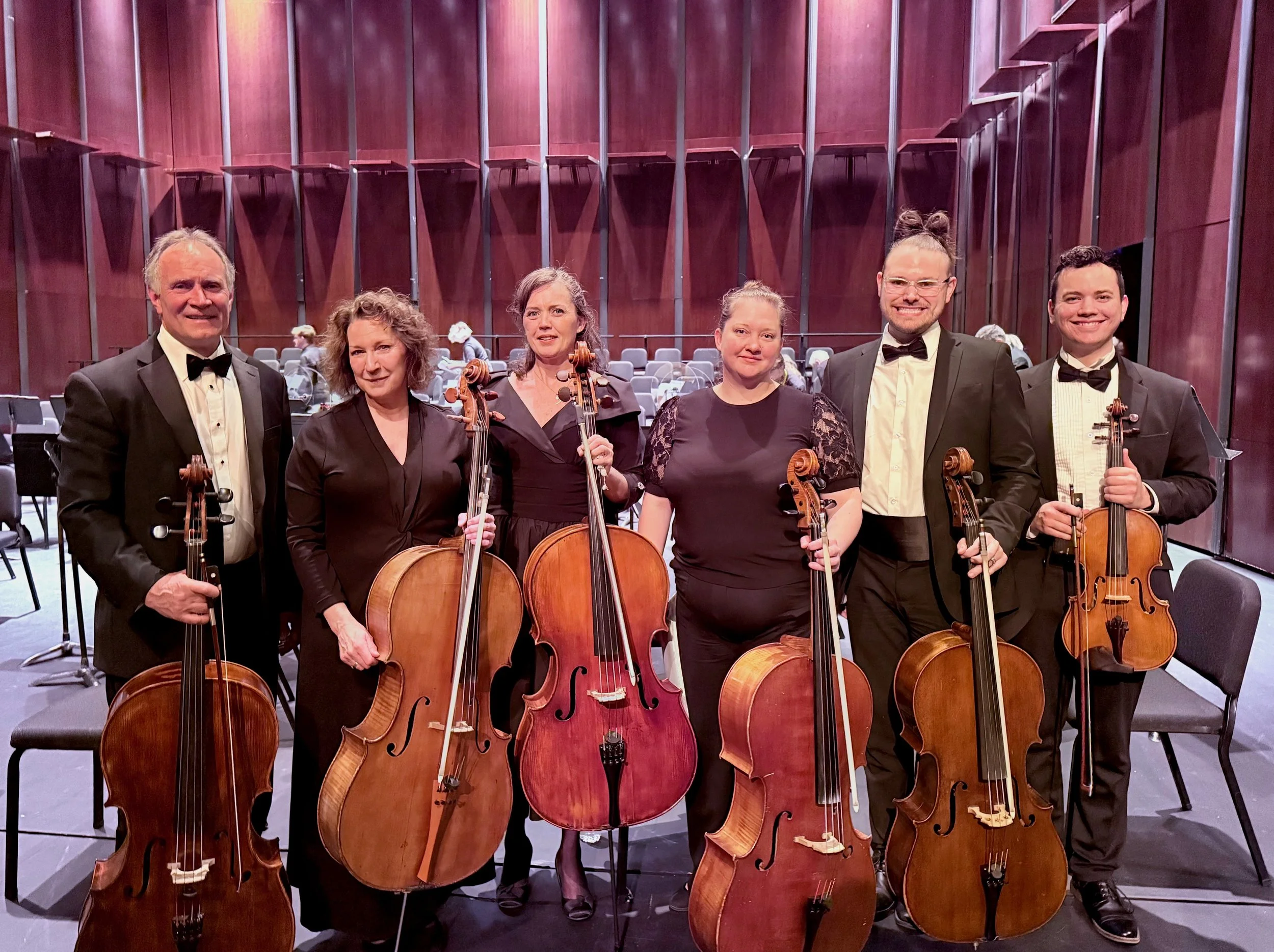 A group of six musicians, four women and two men, dressed in formal black attire, holding string instruments including violins and cellos, inside a concert hall with empty chairs and a wooden stage background.