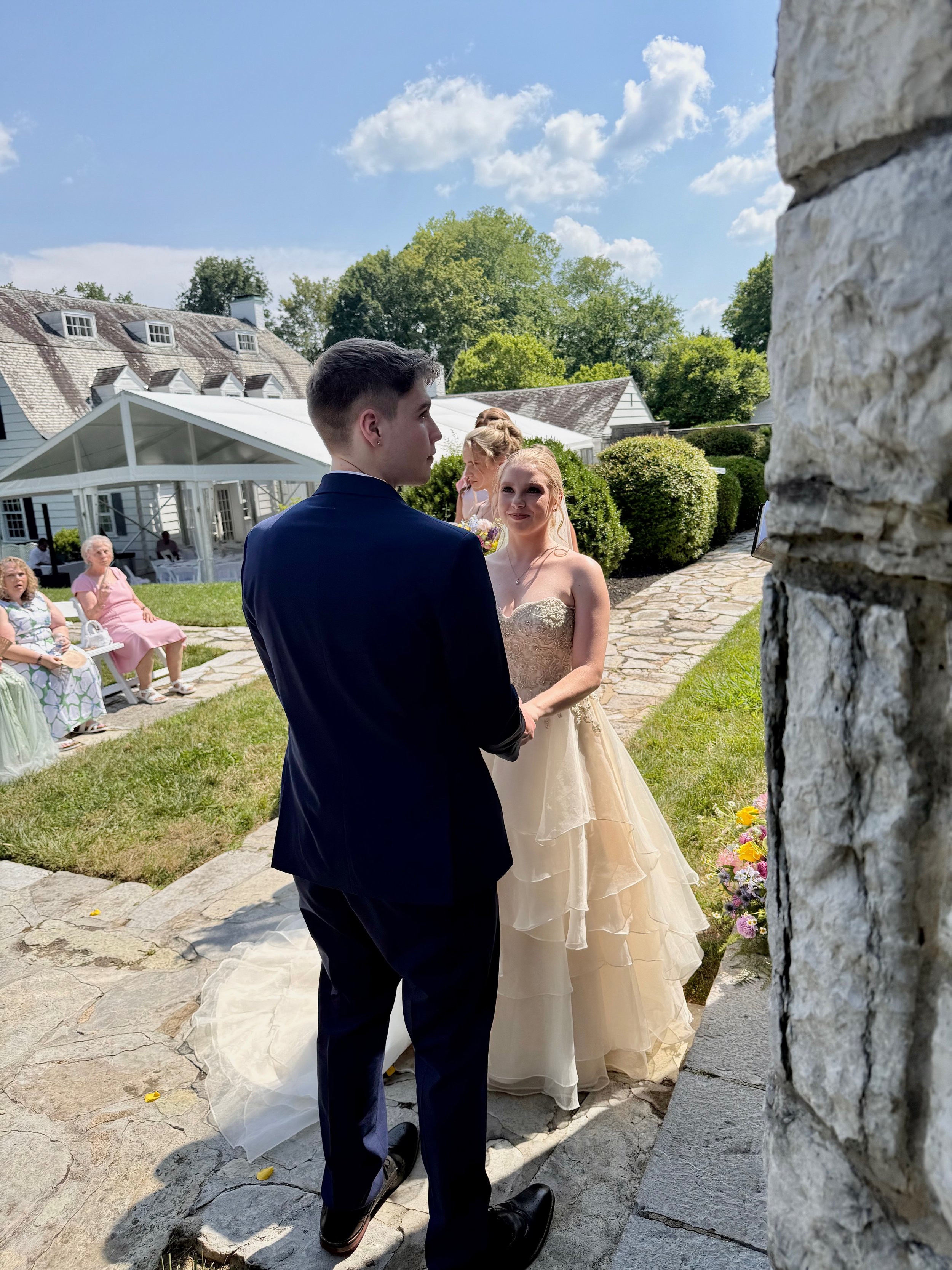 A wedding ceremony outdoors with a bride and groom holding hands, surrounded by guests on a sunny day with blue skies and fluffy clouds.