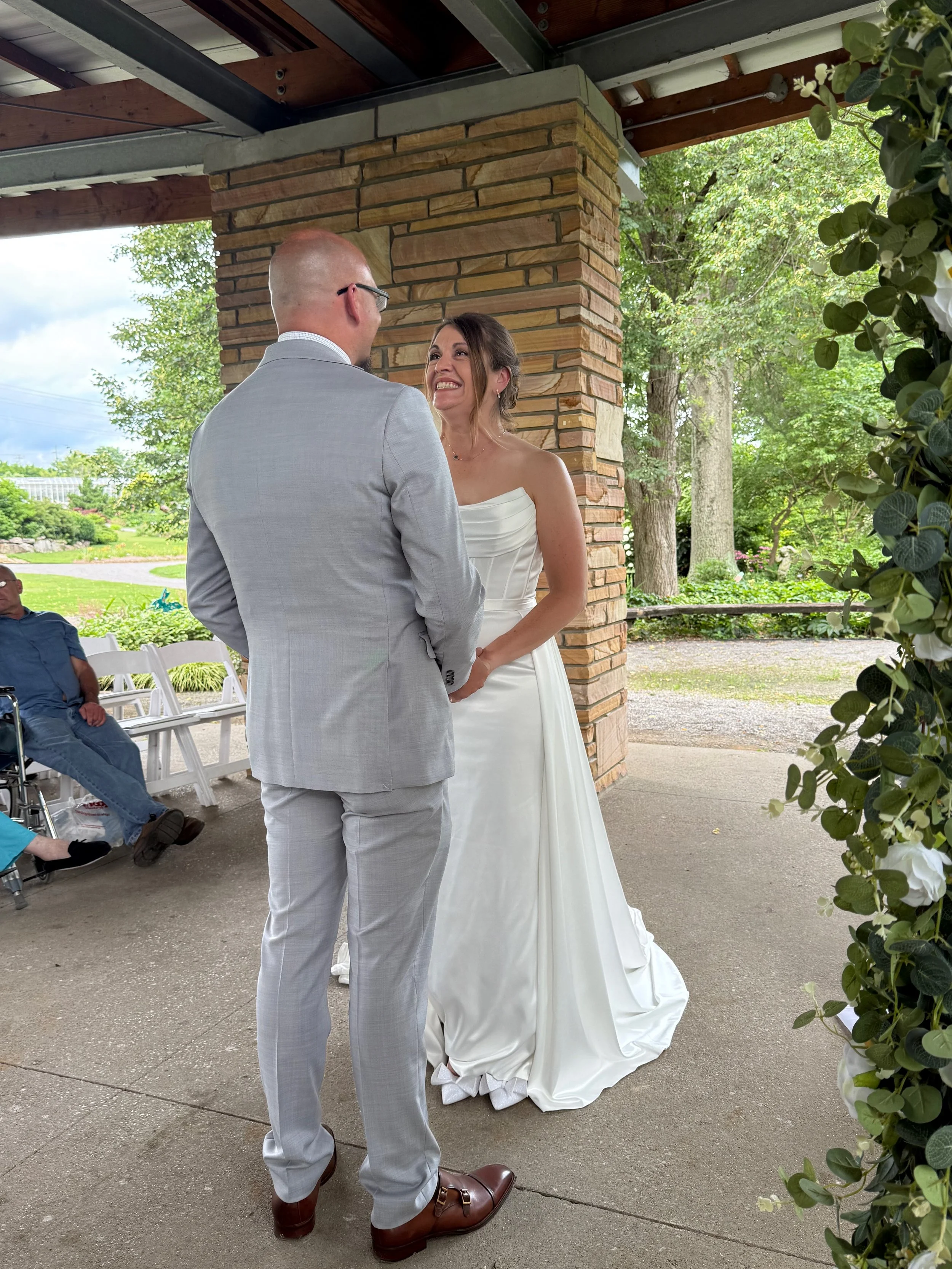 A bride and groom during their wedding ceremony, standing outside under a covered area with greenery in the background. The bride is smiling at the groom, who is facing her. The bride wears a strapless white wedding dress, and the groom wears a light
