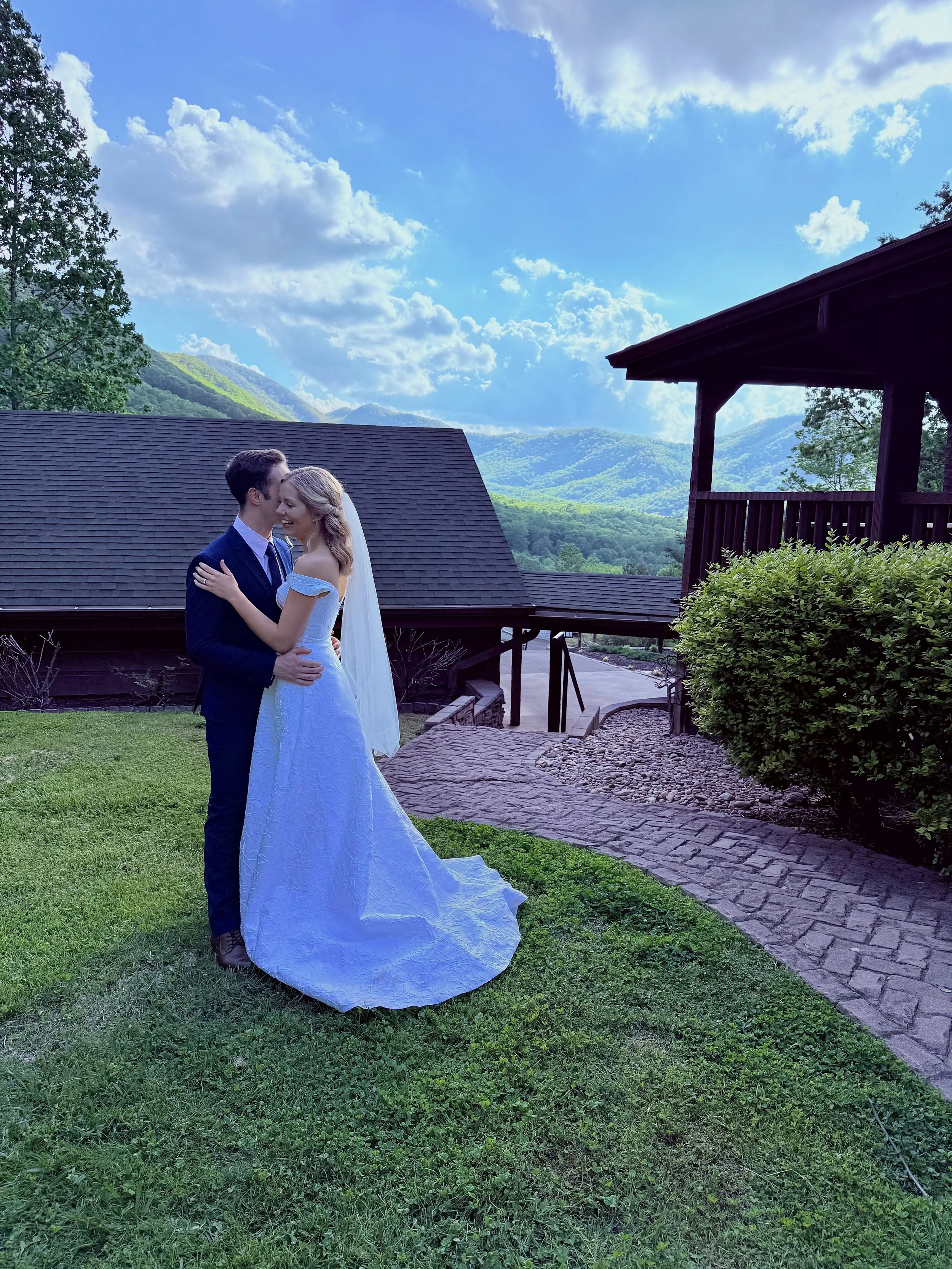 A bride and groom share a dance outdoors on a lush green lawn, with mountains and blue sky with clouds in the background.