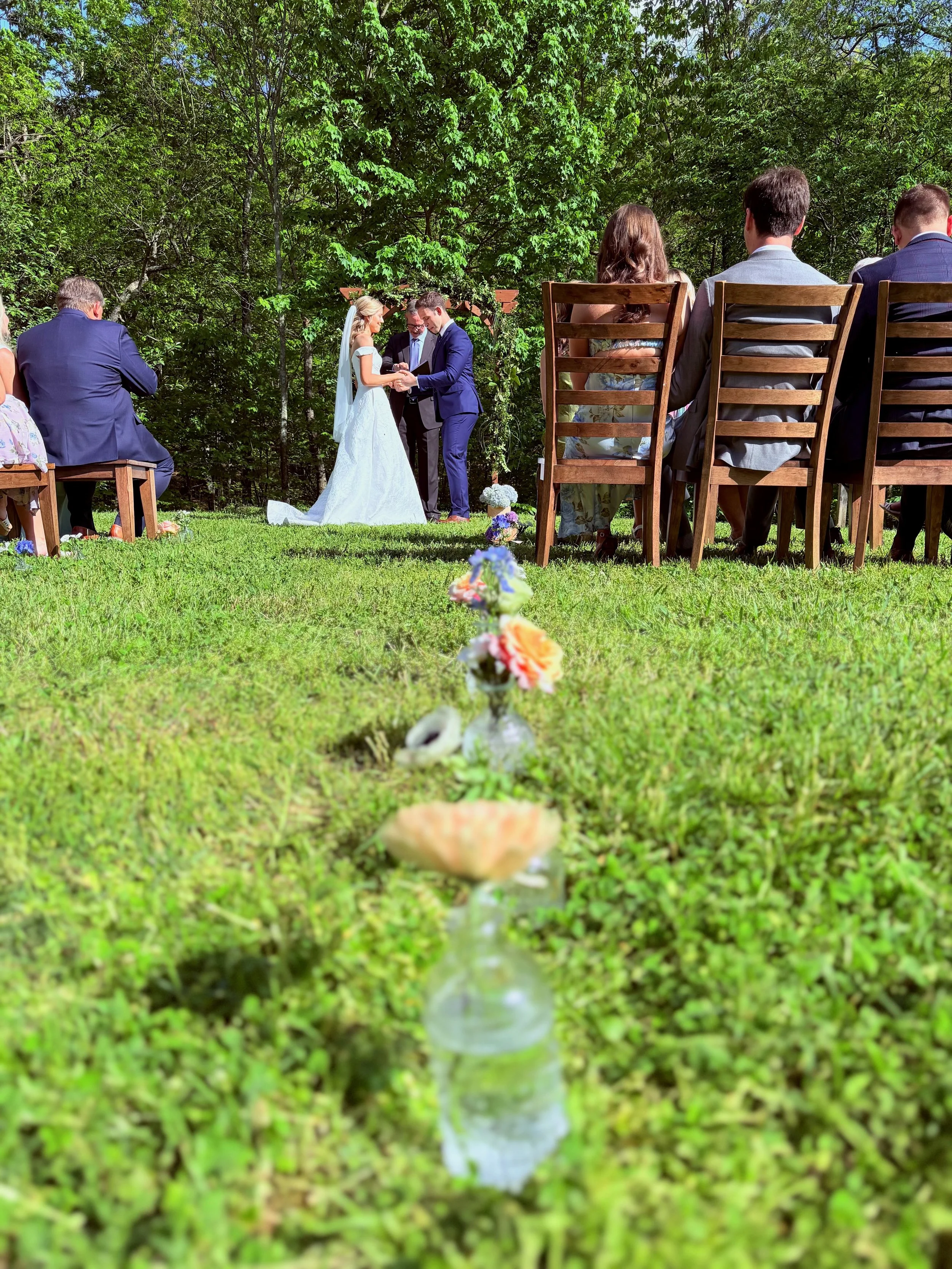 A wedding ceremony taking place outdoors on a grassy area surrounded by trees, with the bride and groom exchanging rings and seated guests watching.