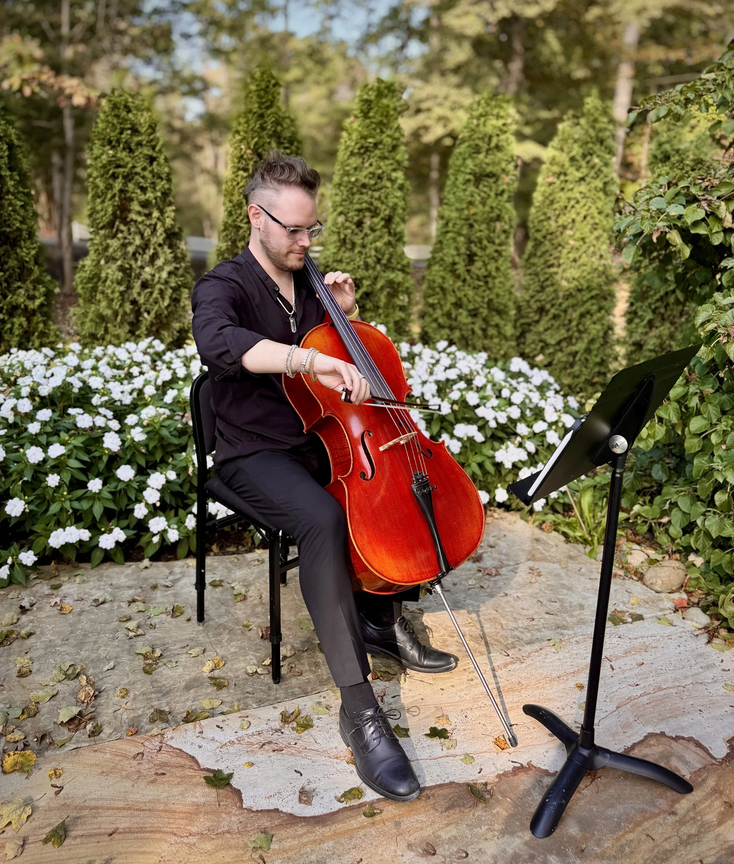 A man playing a cello outdoors, sitting on a black chair with sheet music on a stand in front of him, surrounded by green trees and white flowers, with fallen leaves on the ground.
