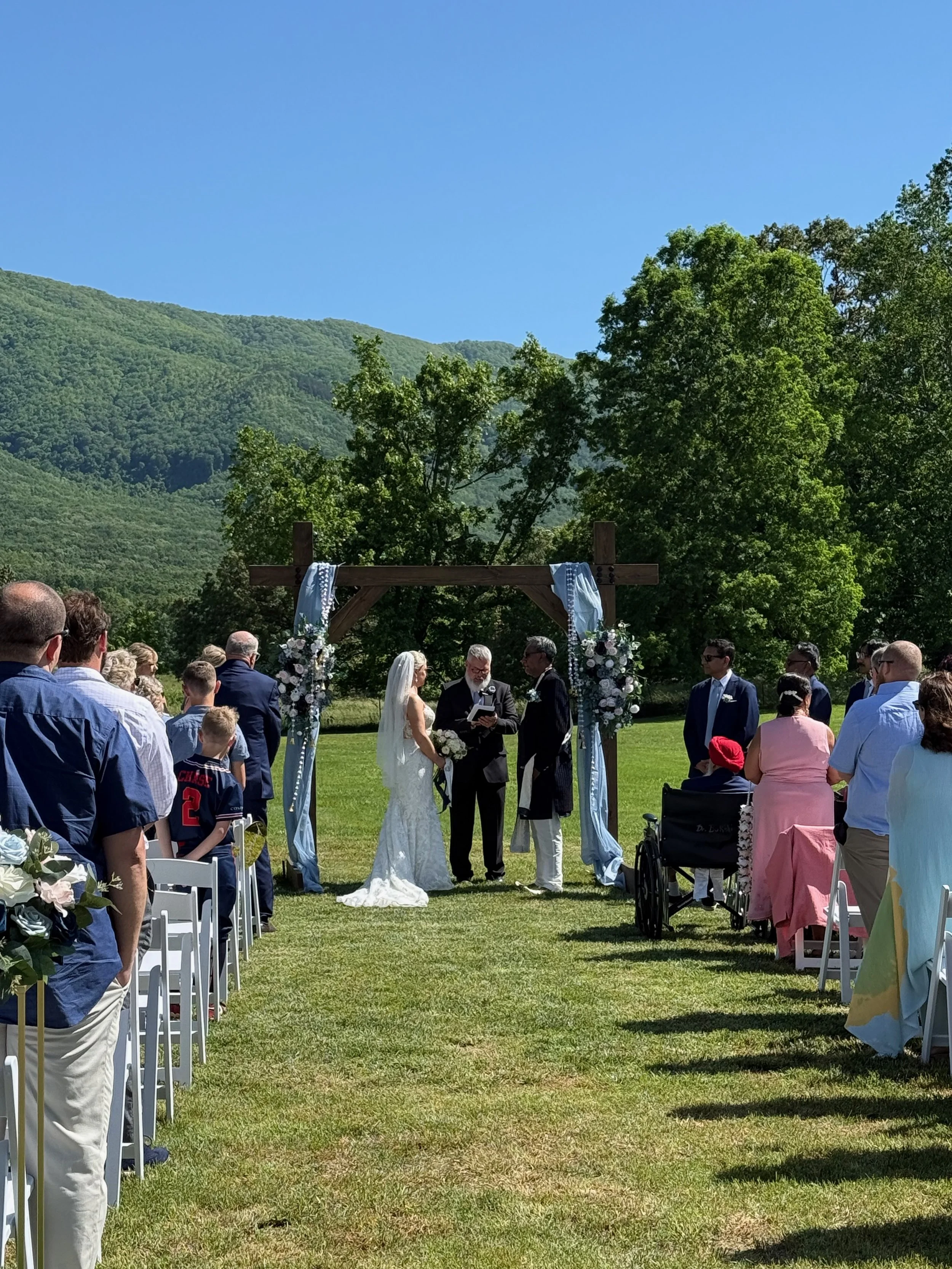 Wedding ceremony outdoors with a couple, officiant, and guests, under a wooden arch decorated with flowers and fabric, set in a green field with mountains and trees in the background.