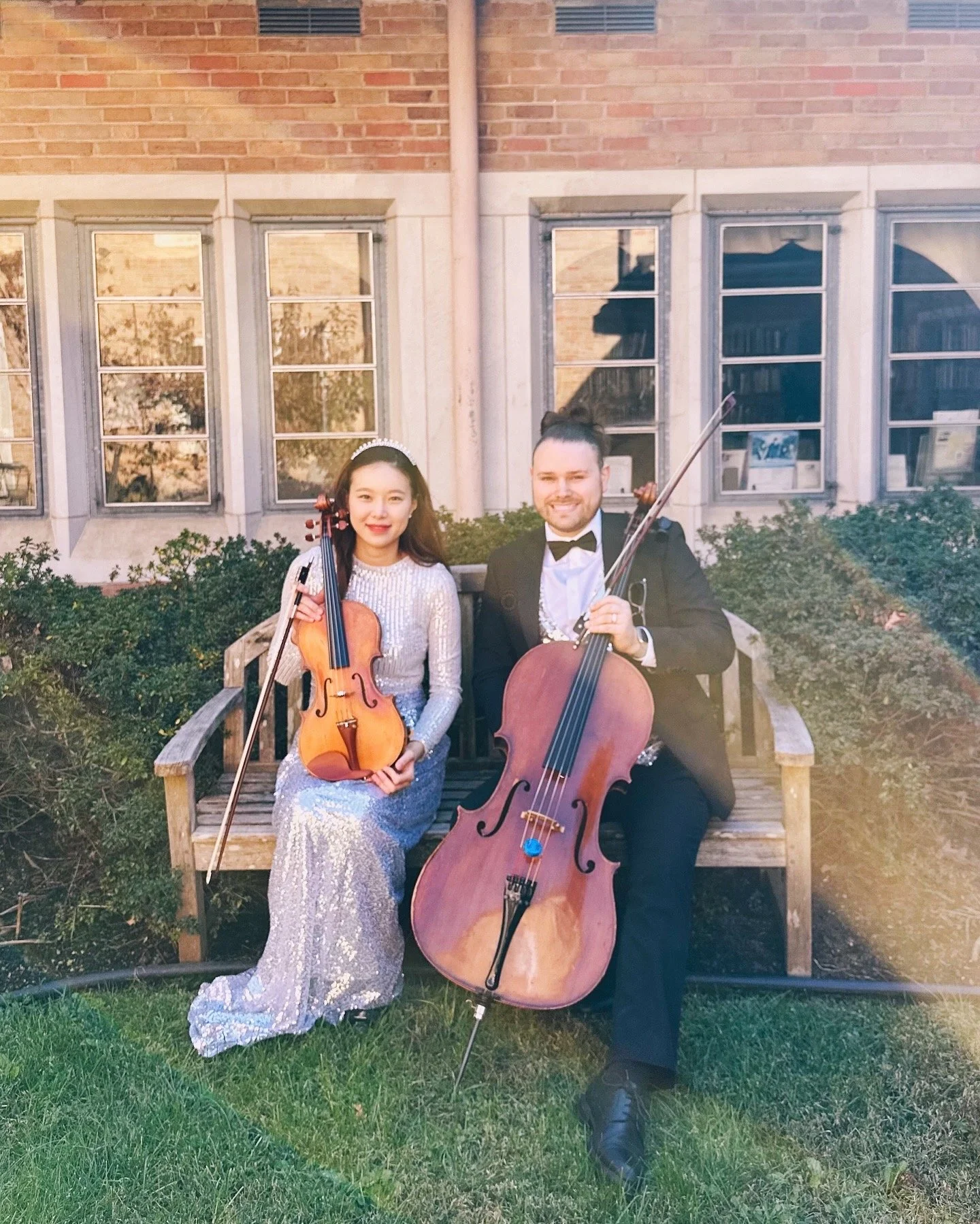 Two musicians, a woman and a man, sitting on a wooden bench outdoors, each holding a string instrument (a violin and a cello), with a brick building in the background.