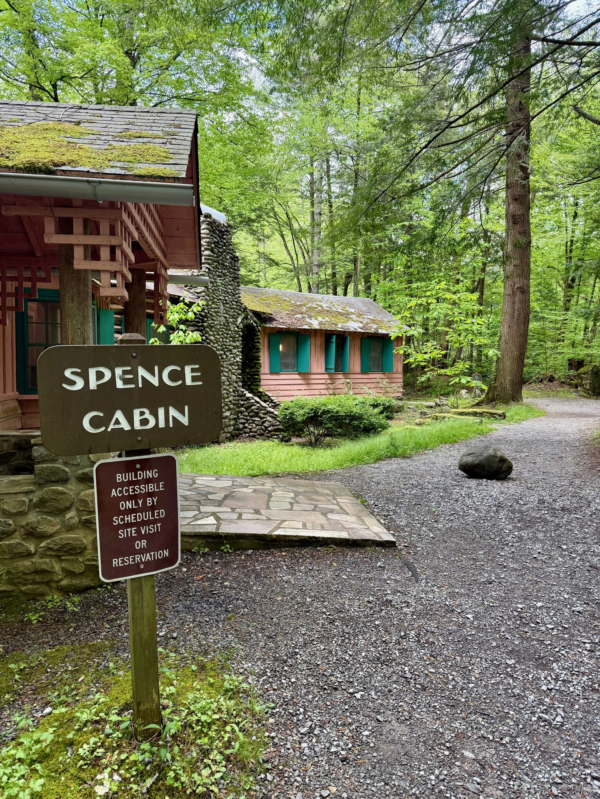A rustic cabin named 'Spence Cabin' in a wooded area with a gravel path and a sign indicating it is accessible only by scheduled site visit or reservation.