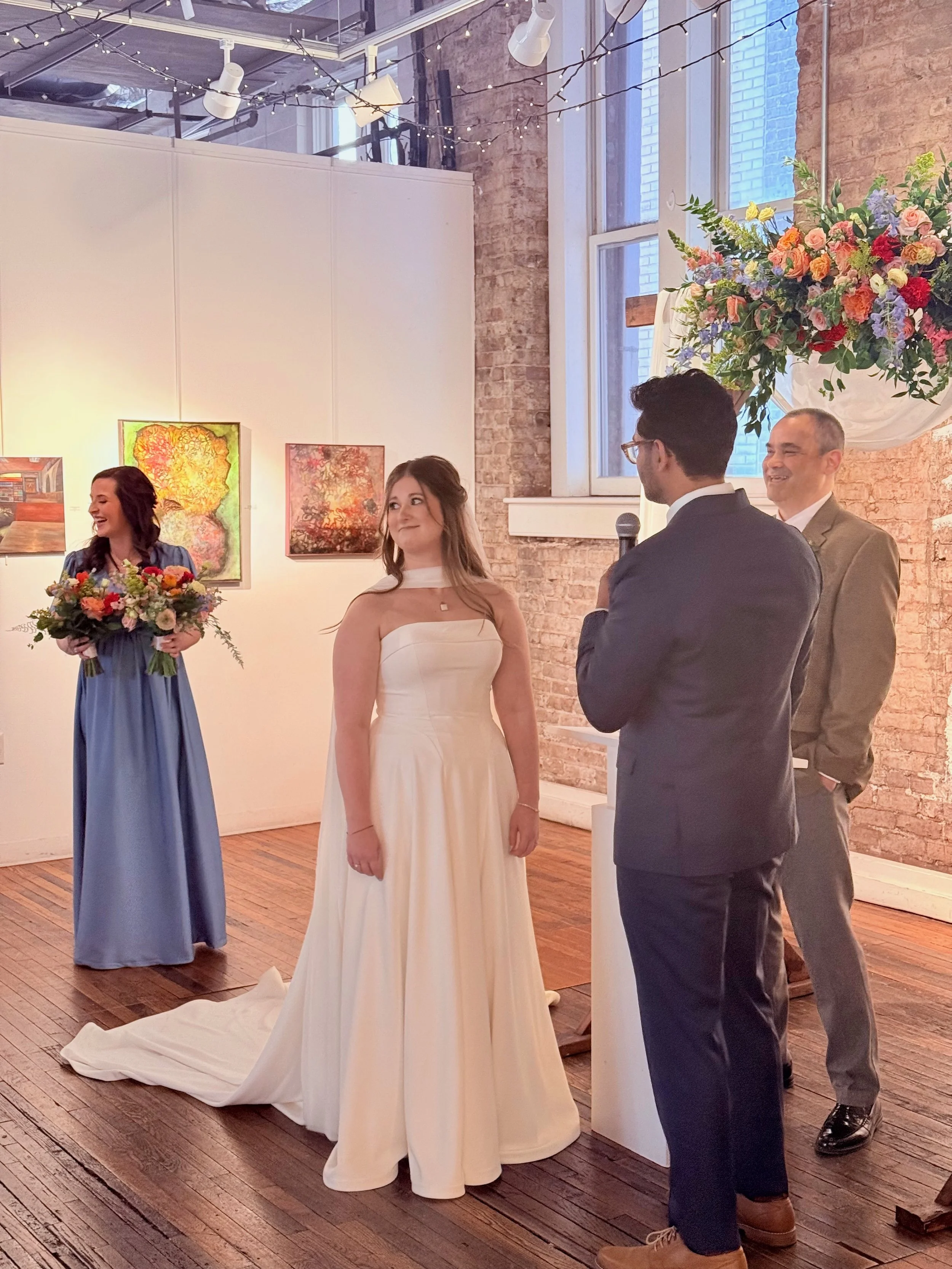 A wedding ceremony in an art gallery with a bride, groom, and officiant, and a bridesmaid holding a bouquet of flowers, with paintings and a large floral arrangement in the background.