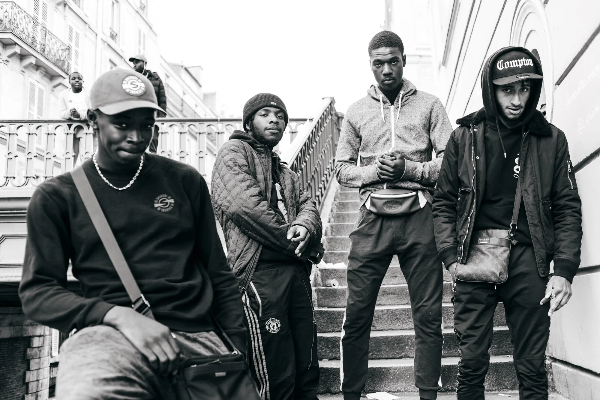 Five young men standing on stairs outdoors in urban setting, dressed in casual streetwear including hoodies, jackets, and caps.