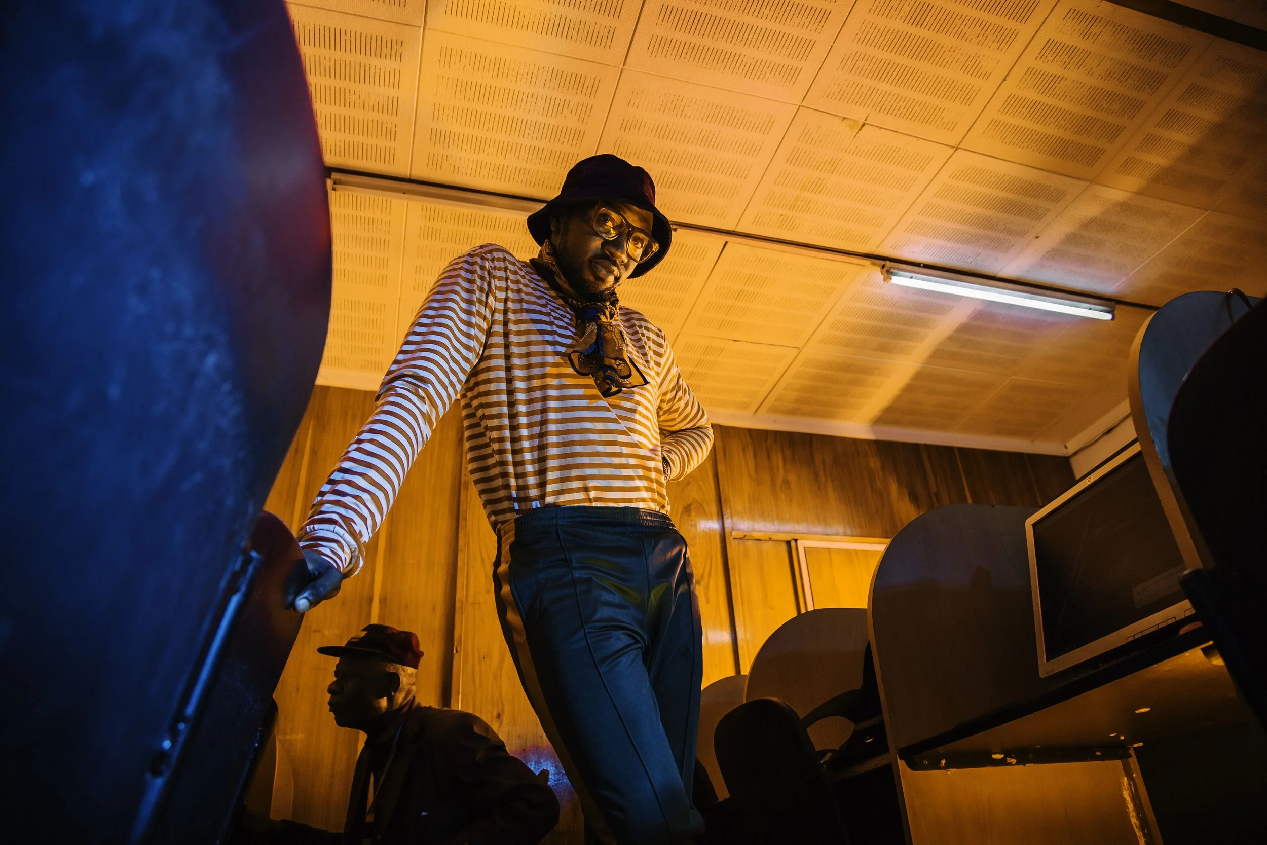 A man wearing a black bucket hat, glasses, a red and white striped long-sleeve shirt, and blue pants stands with one hand on his hip and the other holding a black suitcase in a wood-paneled room illuminated by orange lighting.