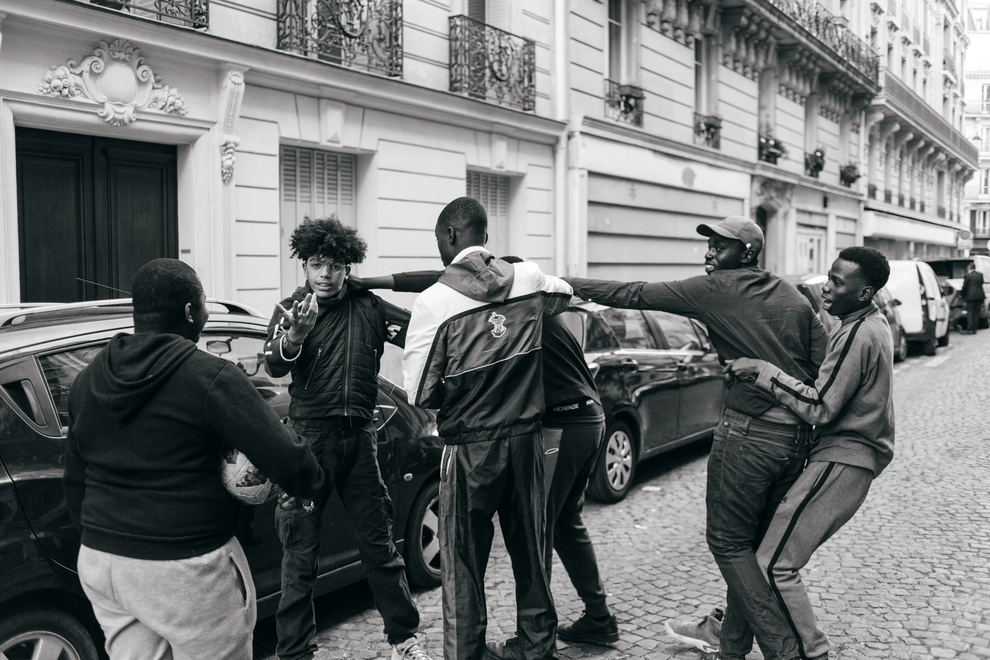 Group of young men engaged in a physical altercation on a city street, with parked cars and buildings in the background.