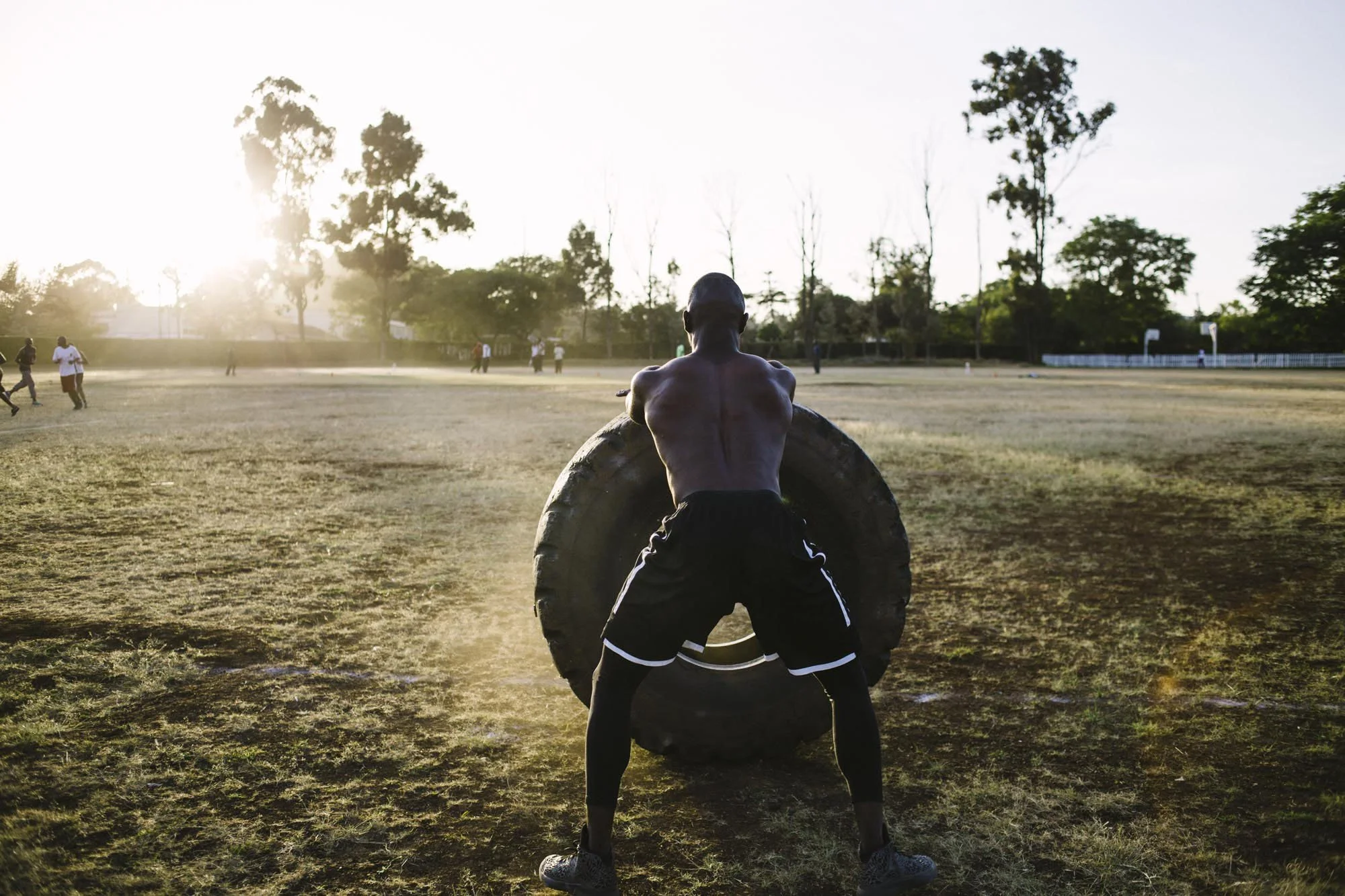 A shirtless man with black shorts and athletic shoes is seen flipping a large tire in a dirt field during sunset with trees and people in the background.