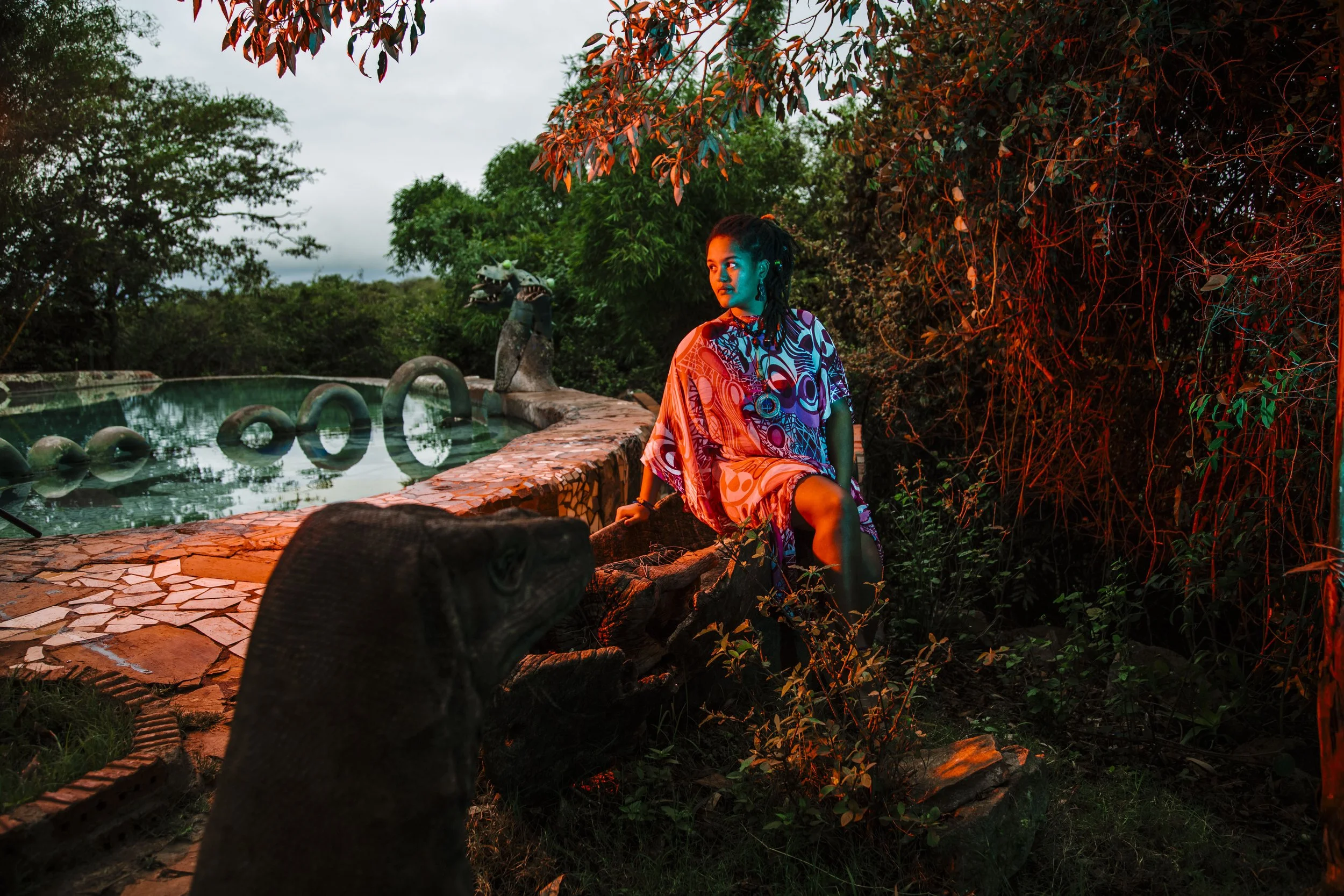 A woman in a colorful patterned dress sitting and leaning against a tree in a lush outdoor setting with trees and bushes, a swimming pool with a dragon sculpture, surrounded by stone and mosaic tiles, with colorful lighting giving a dramatic atmosphe