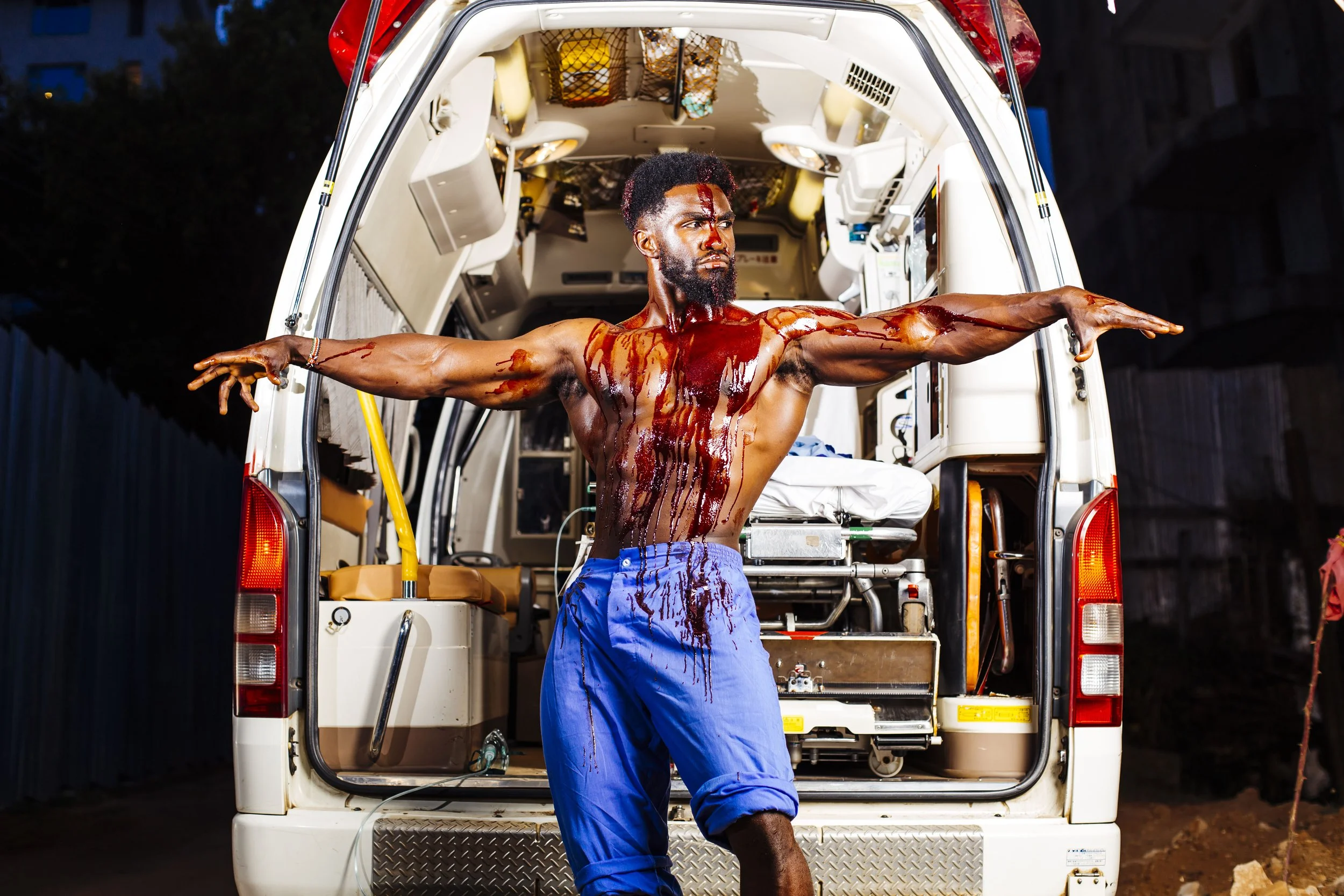 A man with blood on his chest and on his arms stands with arms outstretched in front of an ambulance at night.